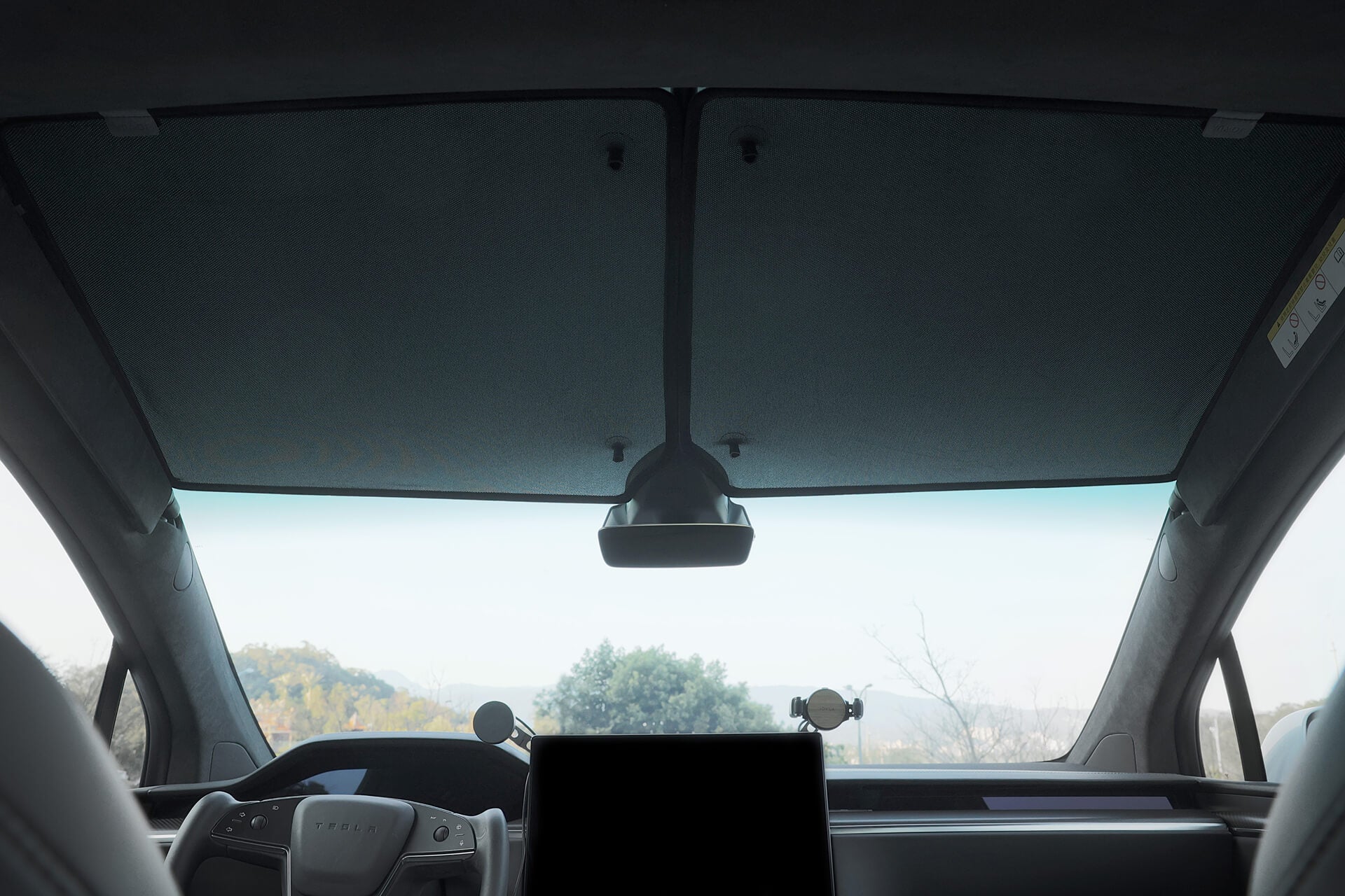 Interior view of a car showcasing the windshield, rearview mirror, and dashboard with a large central screen. Sunlight is gently filtered through JOWUA's Glass Roof Sunshade-Mesh (Model X), offering scenic views of the sky and treetops along with effective UV protection.