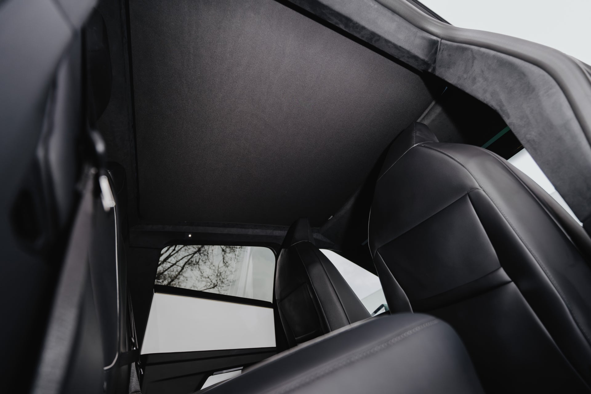 Interior view of a car with JOWUA's Cybertruck Glass Roof Sunshade, showcasing black leather seats and a dark roof lining. The low-angle shot highlights the spaciousness while sunshade clips offer UV protection, and trees are slightly visible outside the window.