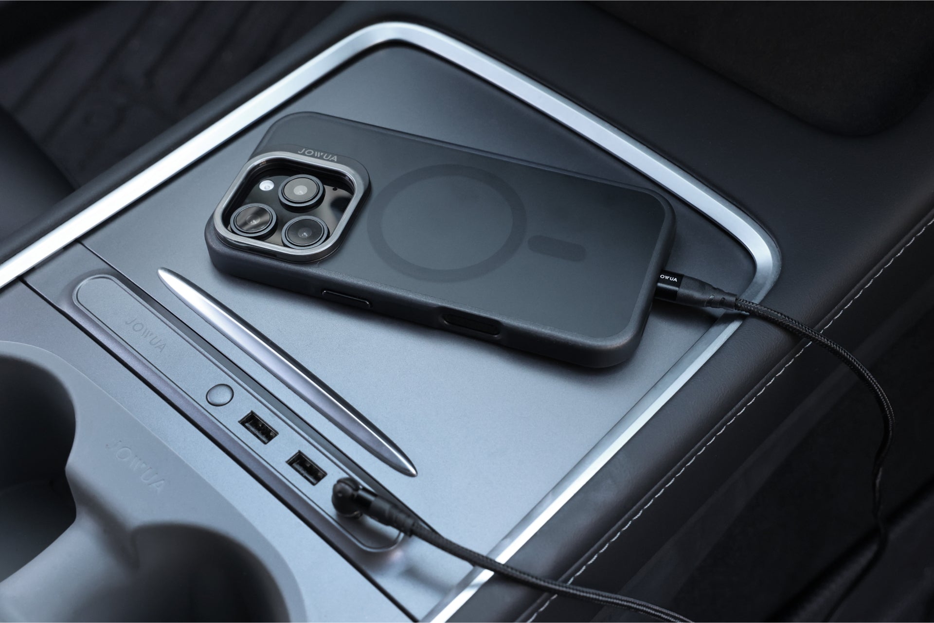 An iPhone 16, protected by a dark JOWUA case designed for Tesla car mounts with MagSafe compatibility and a phone tether tab set, is charging on the car's center console. The console's minimalist design includes USB ports and cup holders. The phone is secured with a black cable for efficient charging while you drive.