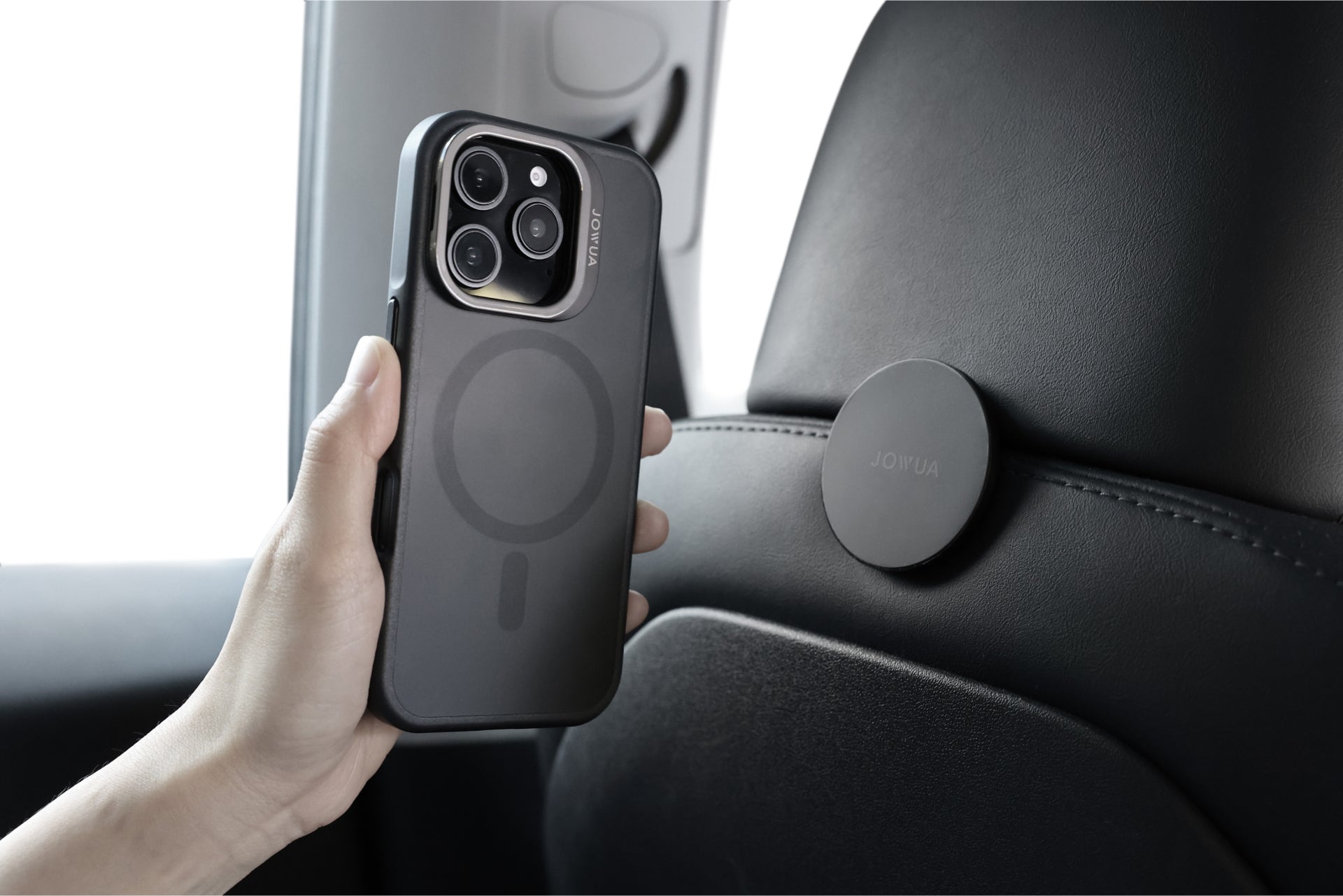 A hand is holding an iPhone 16 encased in a JOWUA iPhone Case designed for Tesla Car Mount, featuring rugged protection and a circular pattern on the back. The phone is positioned near a round magnetic mount on a car seat's headrest, utilizing its MagSafe feature to ensure secure placement.