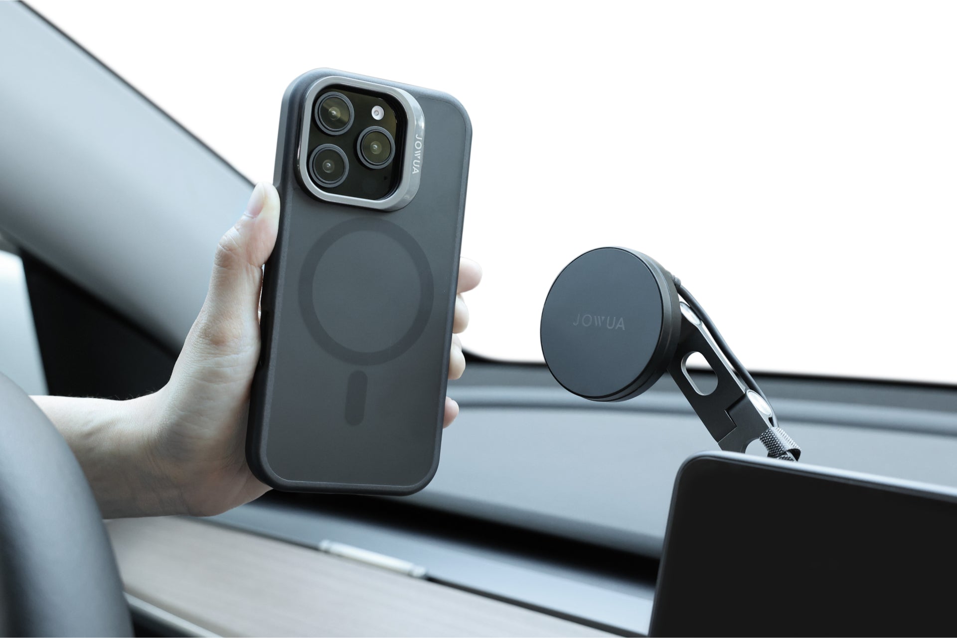 Inside a car, a hand holds an iPhone equipped with the JOWUA iPhone Case for Tesla Car Mount with MagSafe + Phone Tether Tab Set, designed for the iPhone 16 Series. Nearby on the dashboard, a magnetic phone holder stands ready. The bright background accentuates both the phone and holder, perfectly prepared for any journey.