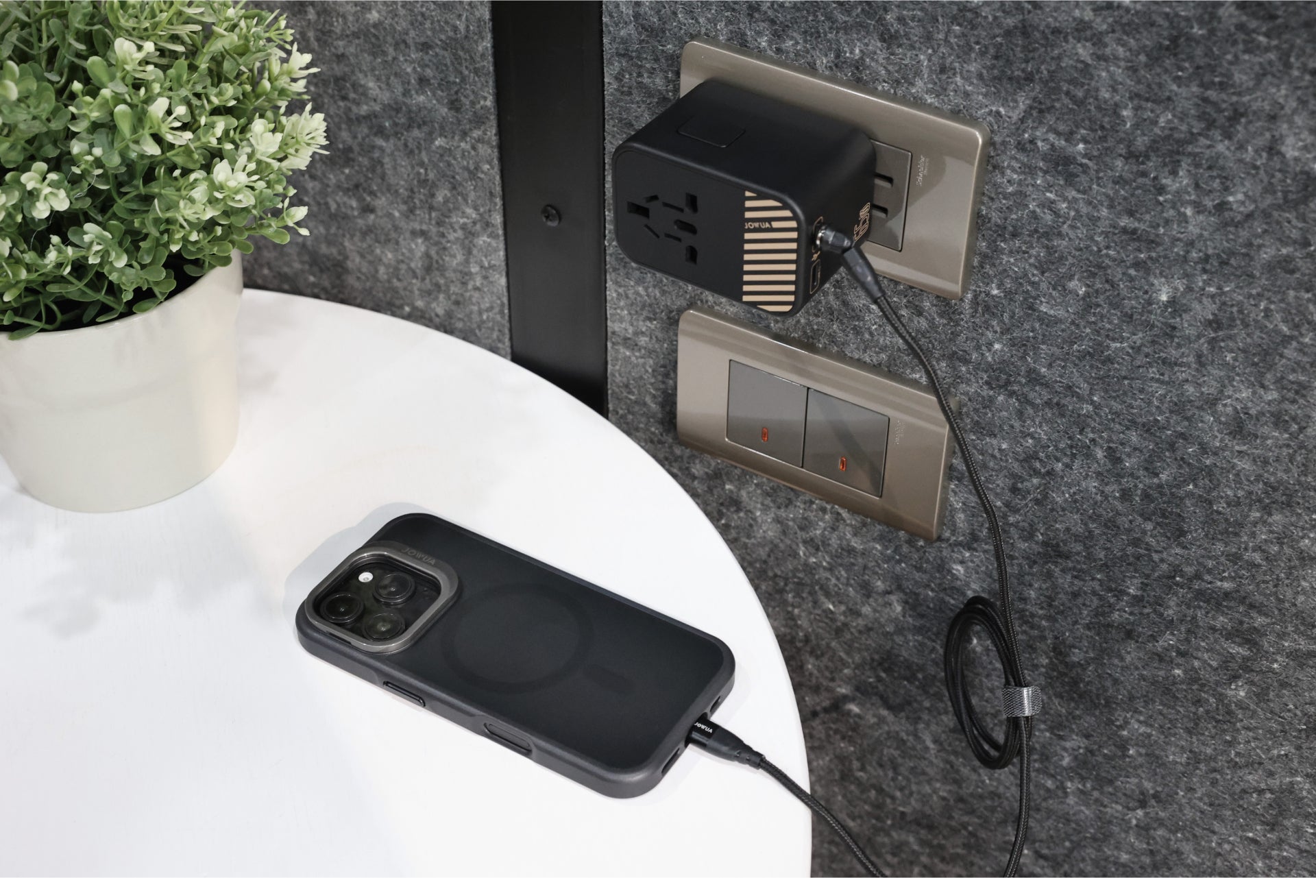 An iPhone 16, equipped with a black JOWUA iPhone Case for Tesla Car Mount with MagSafe and Phone Tether Tab Set, is charging on a white table. The USB cable connects to a power adapter plugged into the wall outlet. A small green plant in a pot is situated on the left side of the table.