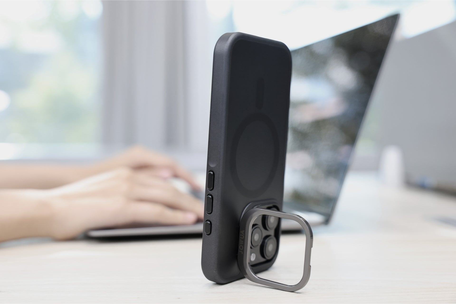 An iPhone 16 Series equipped with a JOWUA iPhone Case for Tesla Car Mount featuring a MagSafe ring stand rests upright on a table. In the background, someone is typing on a laptop, and the environment resembles a clean, brightly lit workspace.