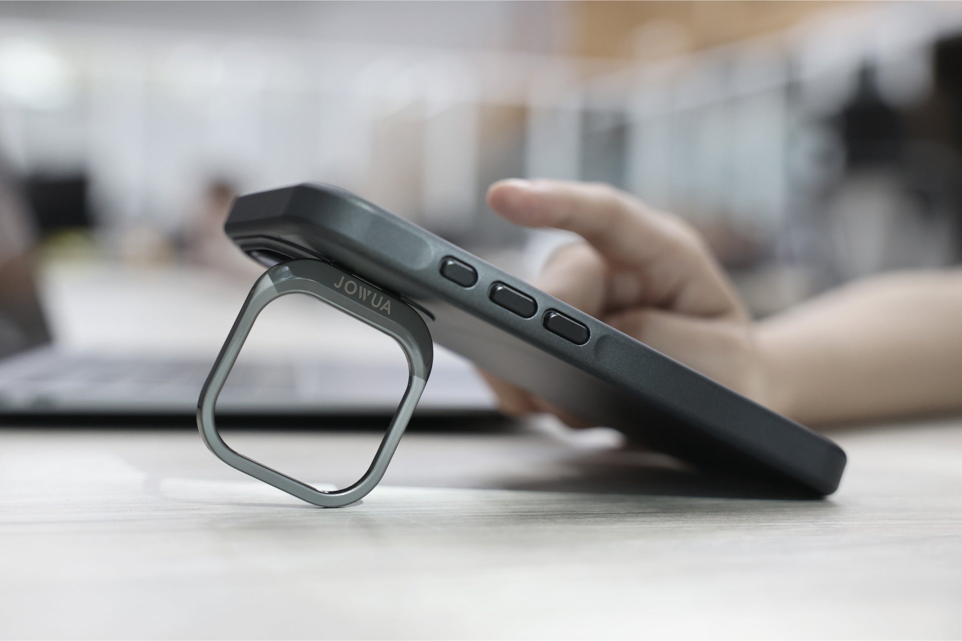 A person places an iPhone in a JOWUA iPhone Case for Tesla Car Mount with MagSafe + Phone Tether Tab Set (iPhone 16 Series) on a table, utilizing the square metal kickstand attached to the back. The phone is angled, revealing three side buttons. In the background, a laptop is partially visible on a blurred desk.