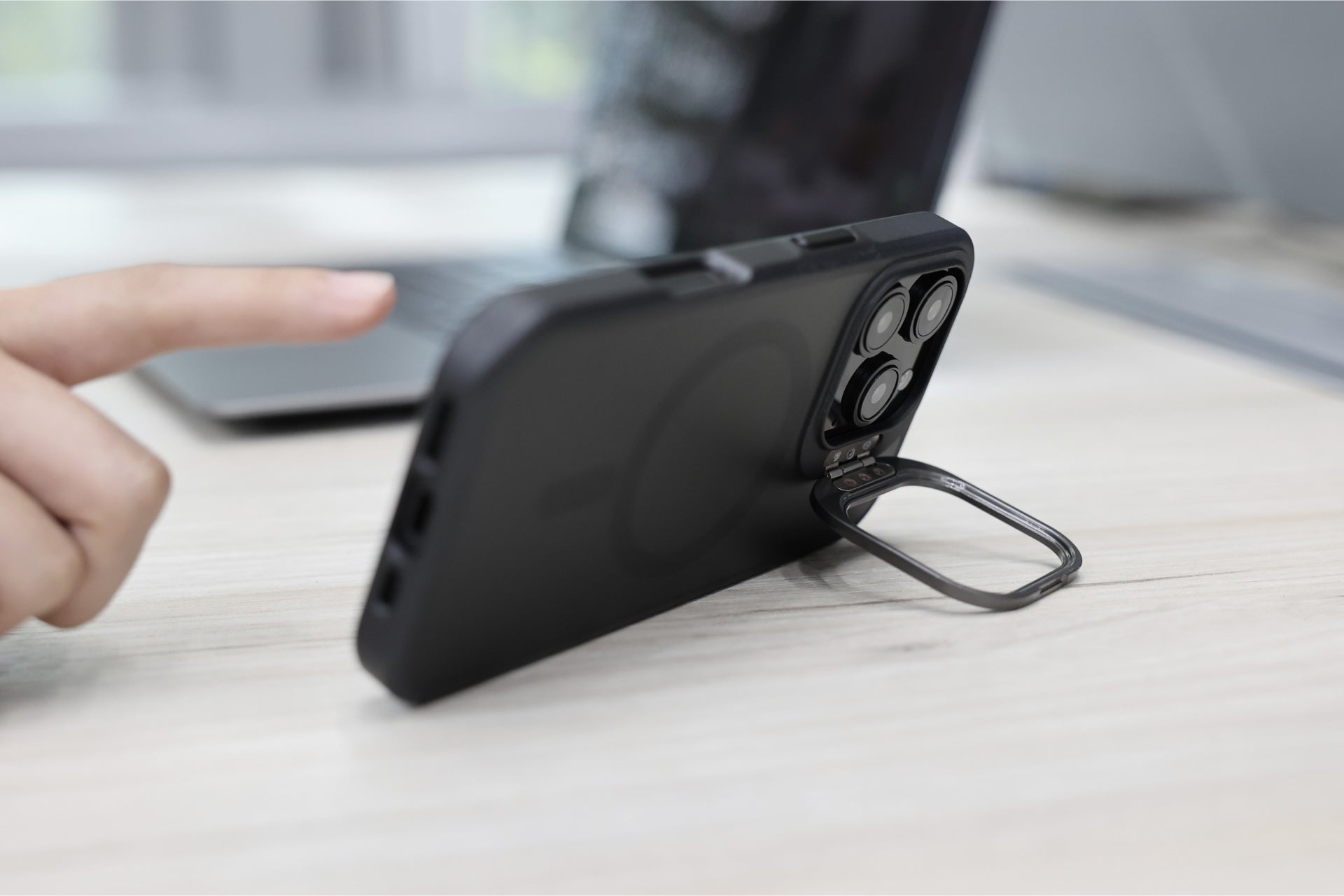 An iPhone 16, encased in a JOWUA iPhone Case for Tesla Car Mount with MagSafe and a Phone Tether Tab Set, is propped on a wooden surface. A finger points towards the clearly focused phone, while a laptop remains blurred in the background.