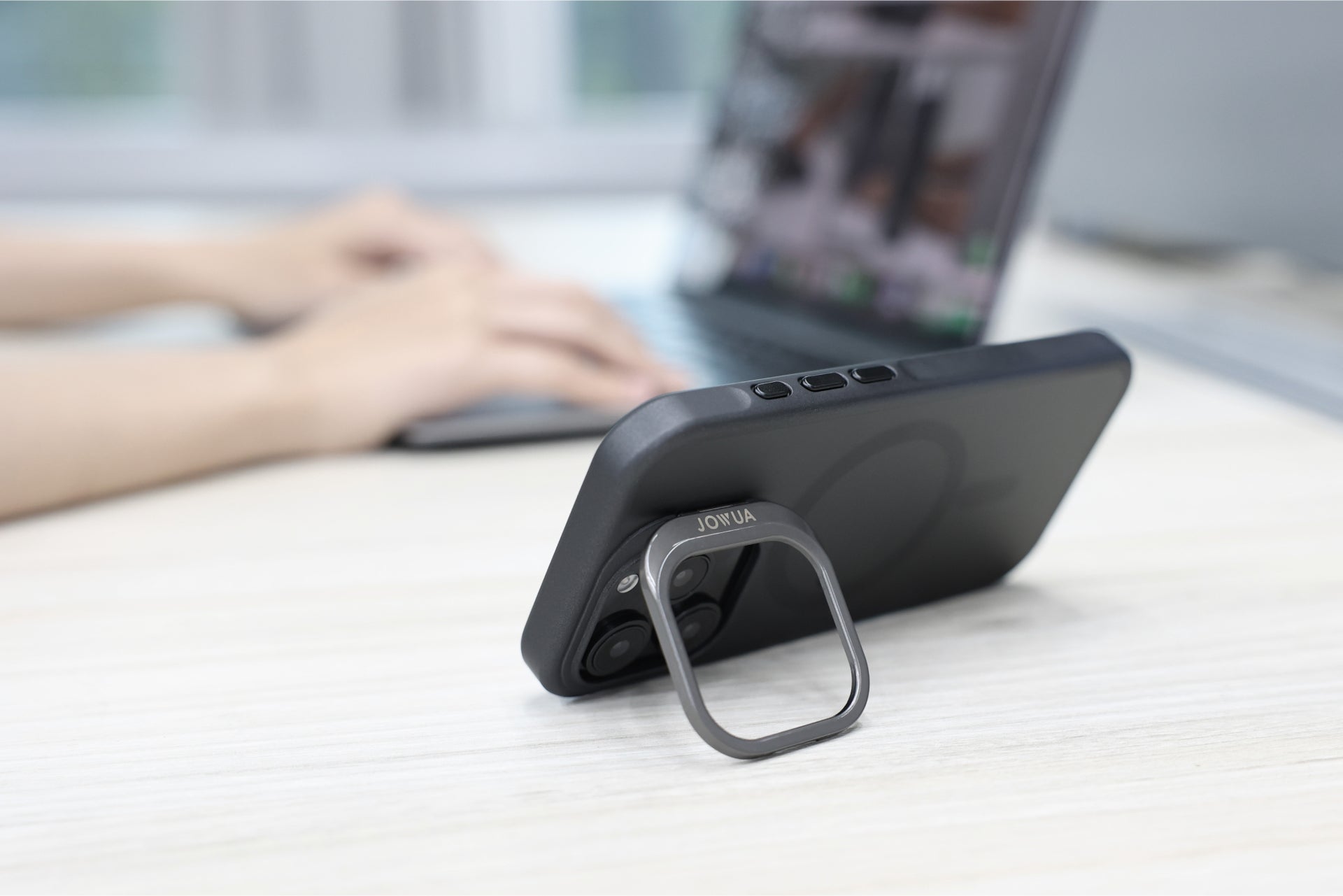 A JOWUA iPhone Case for Tesla Car Mount with MagSafe, featuring a black rugged design and a sleek ring stand, holds an iPhone 16 Series upright on a table. In the background, the blurred hands of someone typing busily on a laptop are visible.