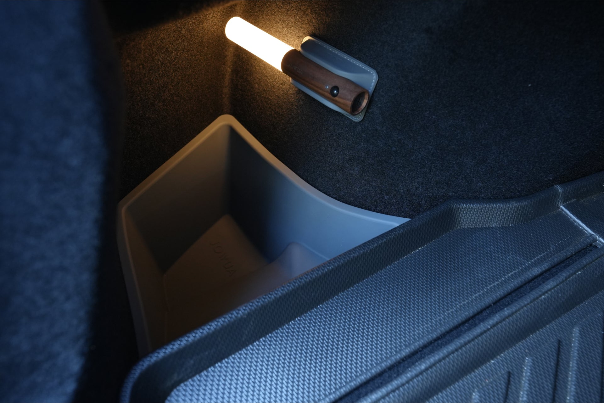 The interior of the car trunk, featuring dark carpet and illuminated by a small corner light, includes a gray plastic bin or organizer. The JOWUA Model 3 All Weather Trunk Combo, specifically reengineered for the 2024+ Model 3 Highland, boasts waterproof TPE material and textured flooring on the right side for both durability and style – an ideal choice for Tesla Model 3 trunk liners.