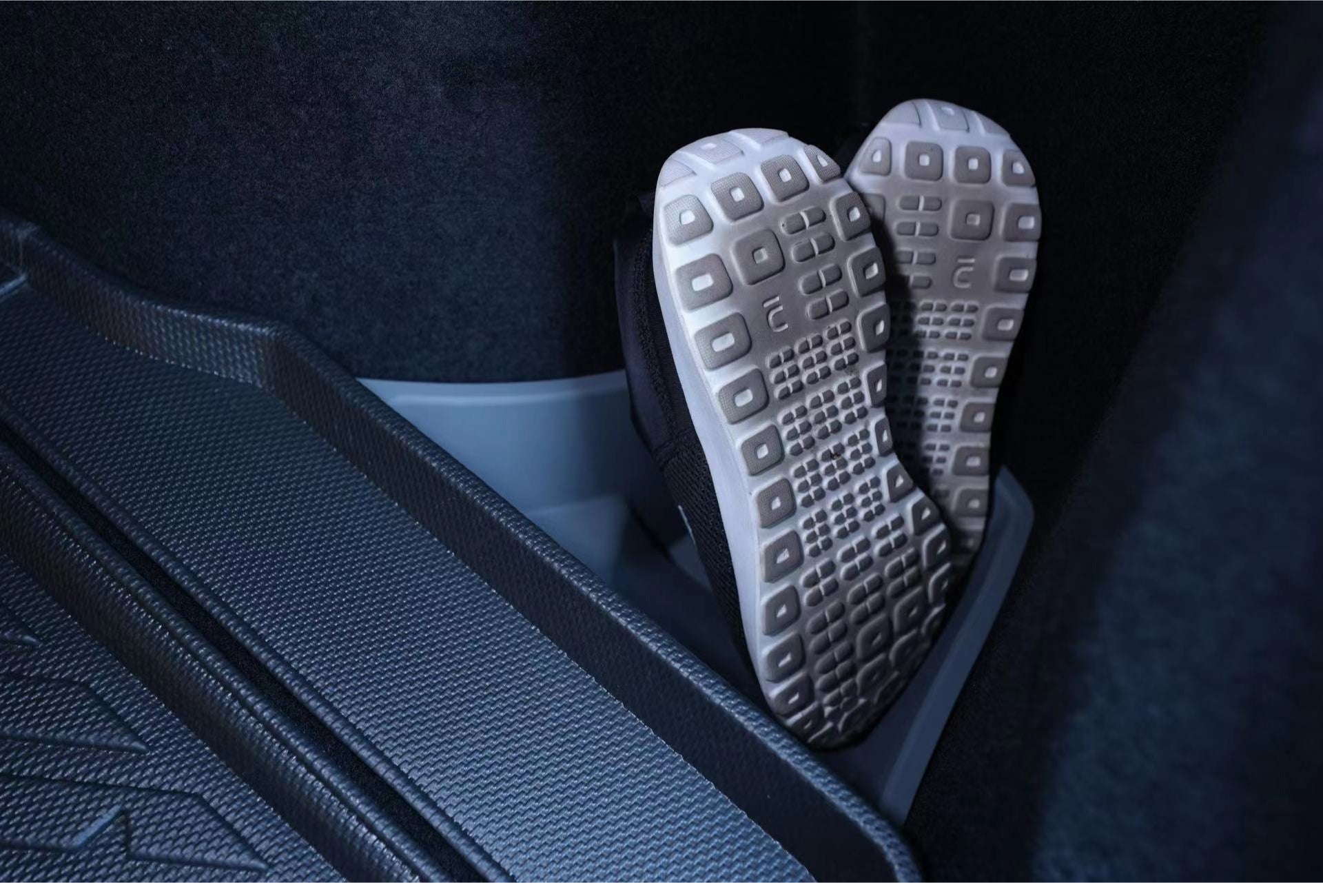 A pair of athletic shoes with light-colored, textured soles is positioned upright in a dimly lit storage area, likely inside the Trunk Storage Bins for Tesla Reengineered Model 3 Highland (2024+) by JOWUA. The compartment features a patterned, non-slip floor surface made from premium silicone.