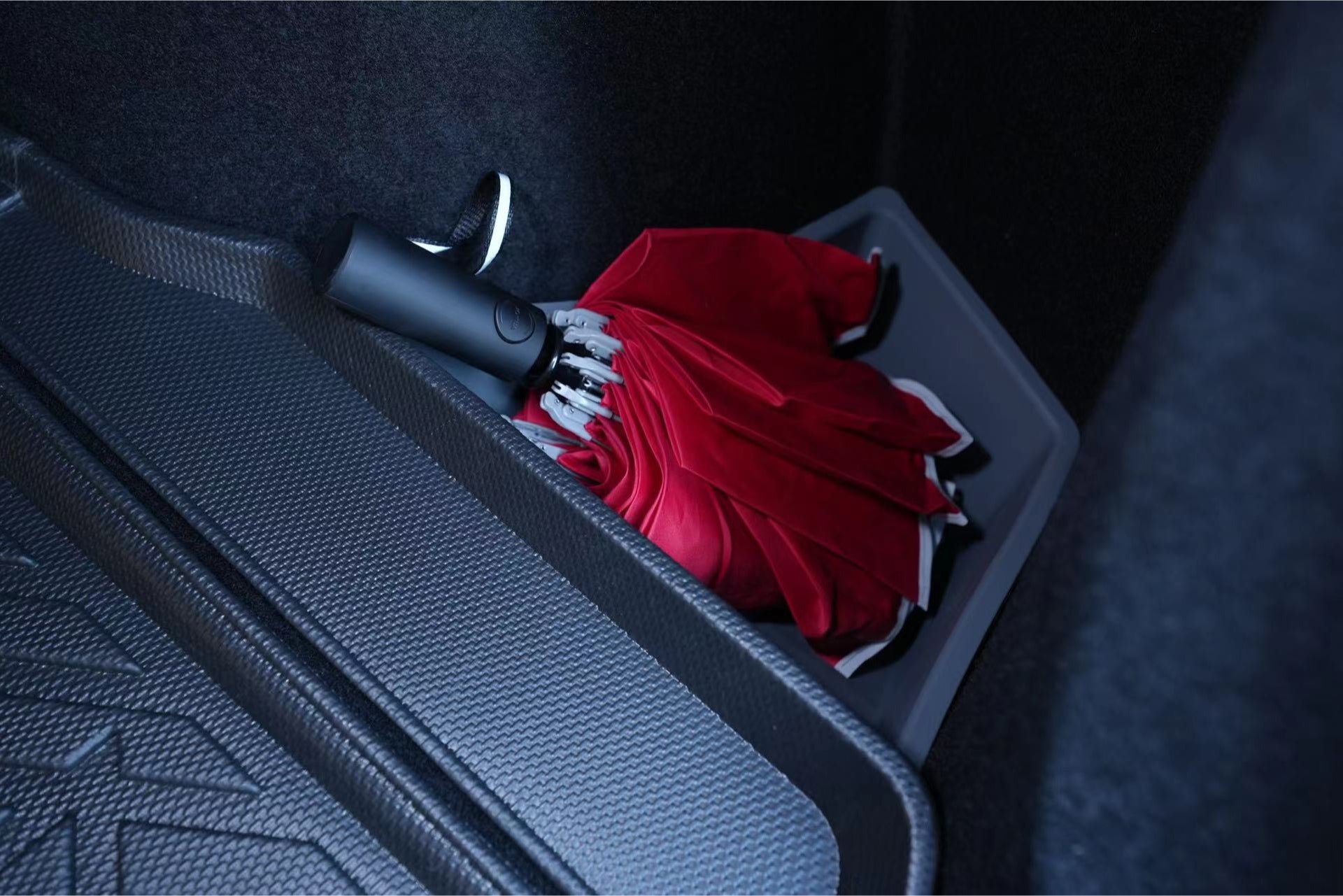 The Model 3 All Weather Trunk Combo from JOWUA stores a red umbrella neatly within a black compartment inside the Tesla Model 3. This compartment features a textured surface, is lined with waterproof TPE material, and blends seamlessly with the car's dark interior.