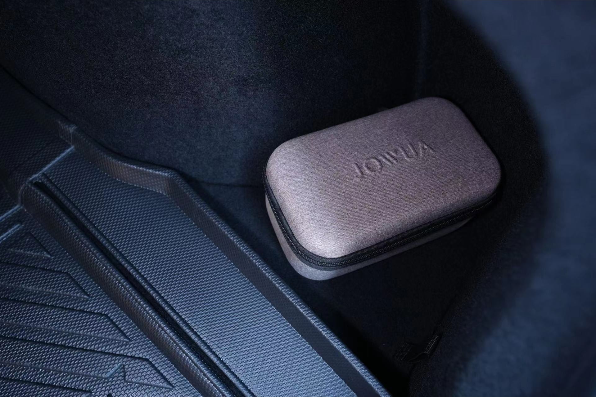 A small, rectangular gray case is nestled beside a textured protective mat in the trunk of the Tesla Reengineered Model 3 Highland (2024+). Crafted from premium silicone, the case features a zipper and elegantly bears the "JOWUA" brand name on top. The trunk's interior is dark yet well-defined.