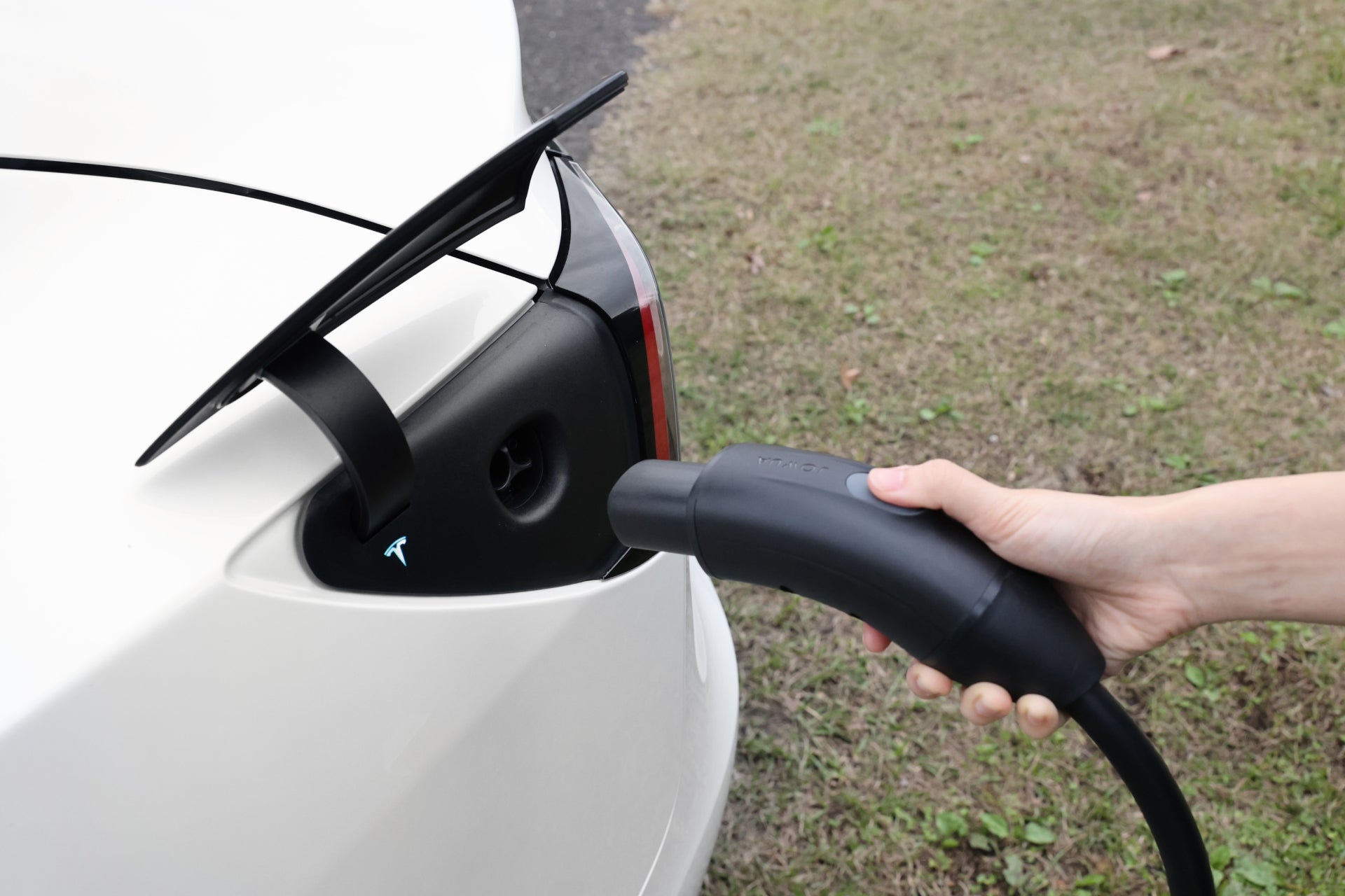 A close-up of a person using the JOWUA NACS Mobile Connector Solution for Tesla to charge an electric car. A hand grips the black charging plug, connecting it to the vehicle's port. The white car is parked outdoors on grassy ground, ready for scheduled charging with its Tesla adapter.