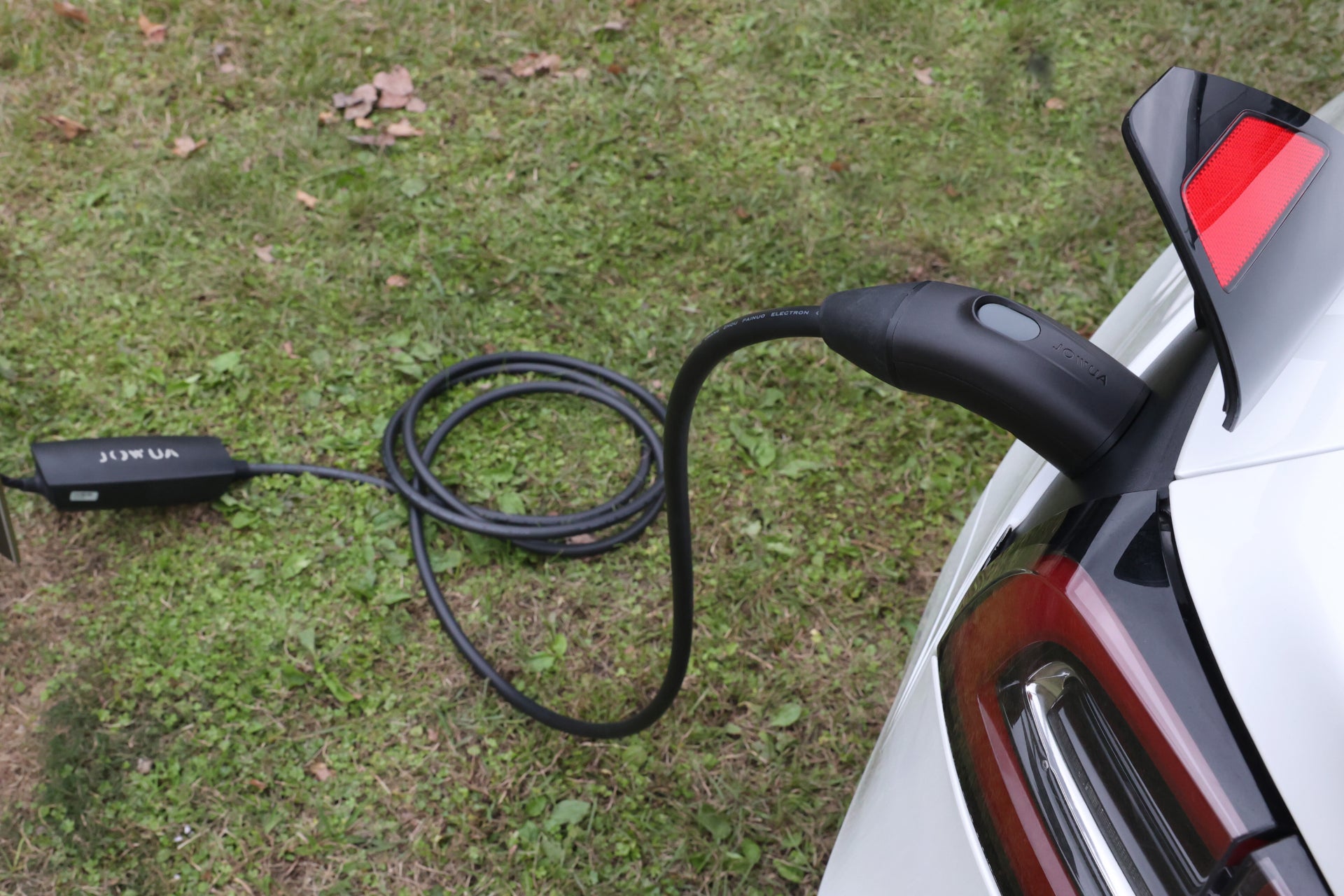 A white electric car sits on a grassy surface, its rear light gleaming softly. The vehicle is connected to the ground-loops of a NACS Mobile Connector Solution for Tesla by JOWUA, ready for scheduled charging.