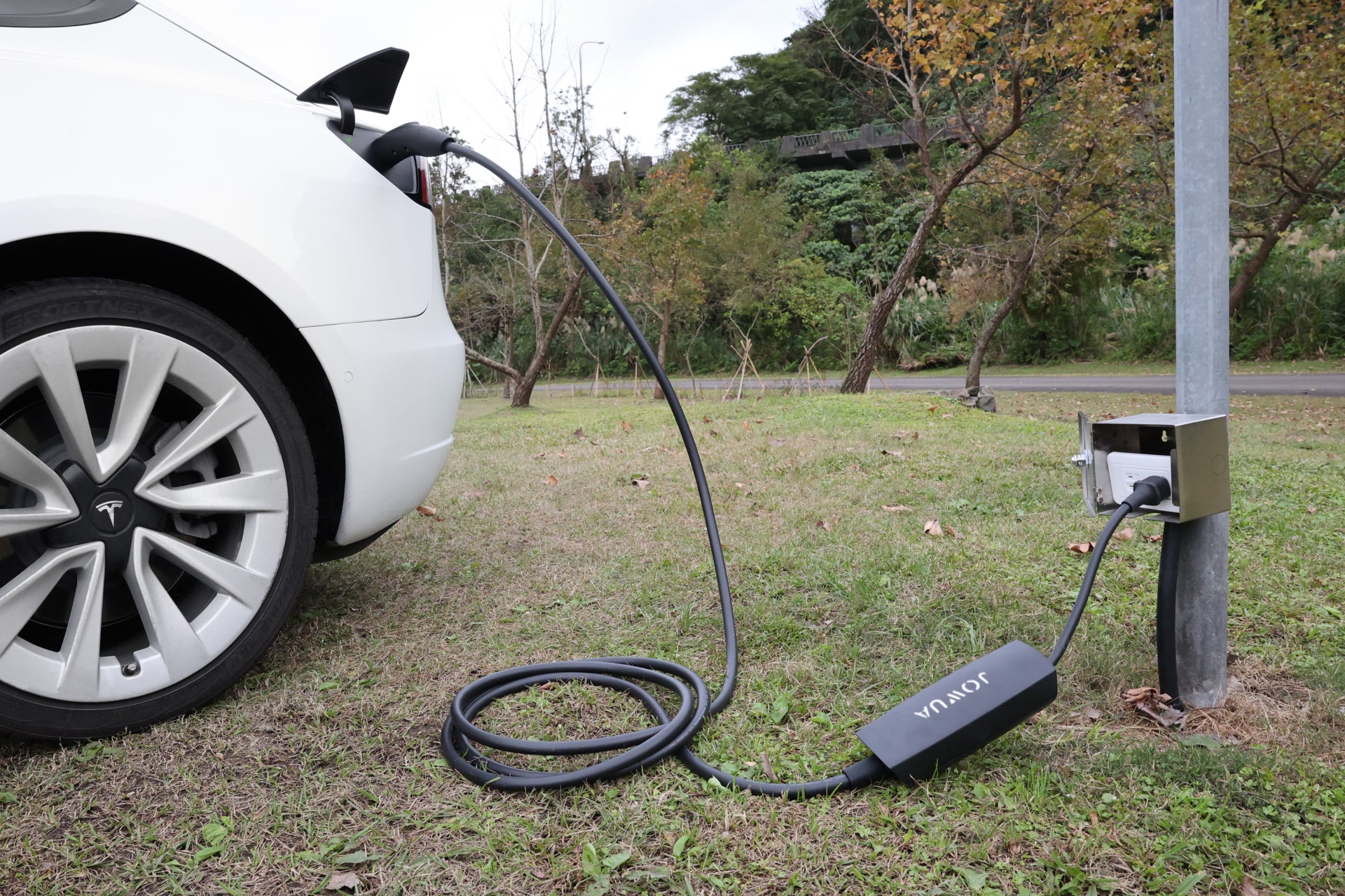 A white electric car is parked on grass and connected to a Tesla via a long cable using the JOWUA NACS Mobile Connector Solution, with a metal pole-mounted charging station framed by trees in the background.