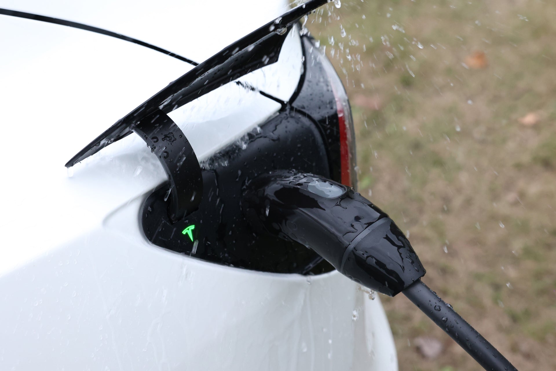 In the rain, a JOWUA NACS Mobile Connector Solution is plugged into the rear port of a white Tesla, with water droplets shimmering on its surface.