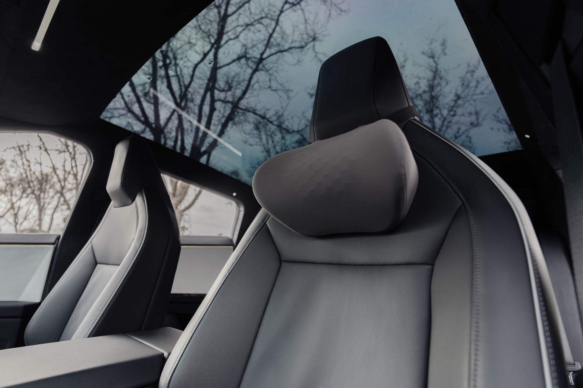 The JOWUA Car Neck Pillow, resting on the ergonomically designed passenger seat, enhances comfort beneath a panoramic glass roof. The seat features dark material with leafless trees visible through the roof, adding a winter ambiance.