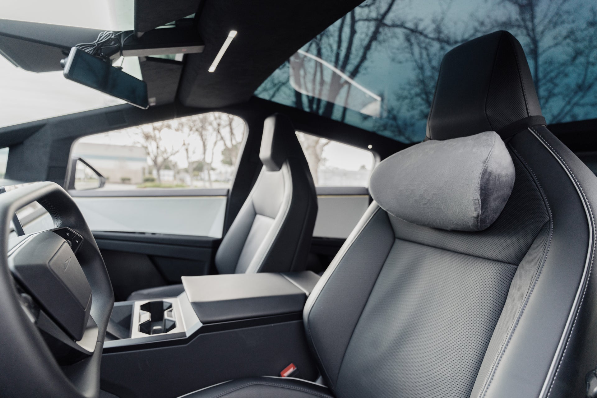 The modern car interior includes two ergonomic black leather front seats, a sleek dashboard, and a large infotainment screen. Above, the glass roof offers views of tree branches against the cloudy sky. Enhance comfort with the JOWUA Car Neck Pillow.