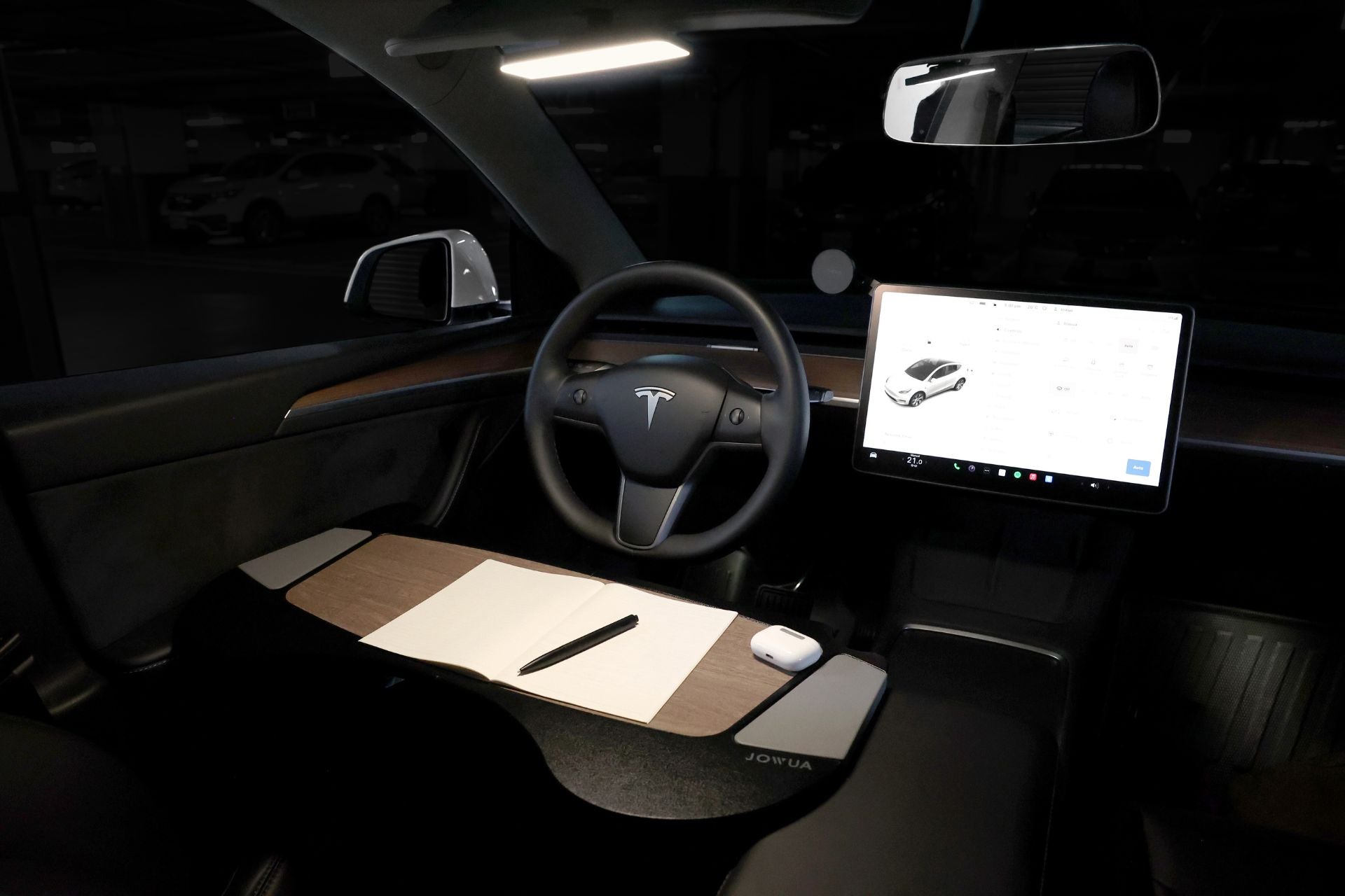 Inside the Tesla car parked in a dimly lit area, the dashboard features a large touchscreen displaying a car interface. Alongside the steering wheel and side mirror, JOWUA's LED Center Console Combo for the Tesla Reengineered Model 3 Highland (2024+) neatly organizes papers and holds a white mouse on the wooden desk surface.