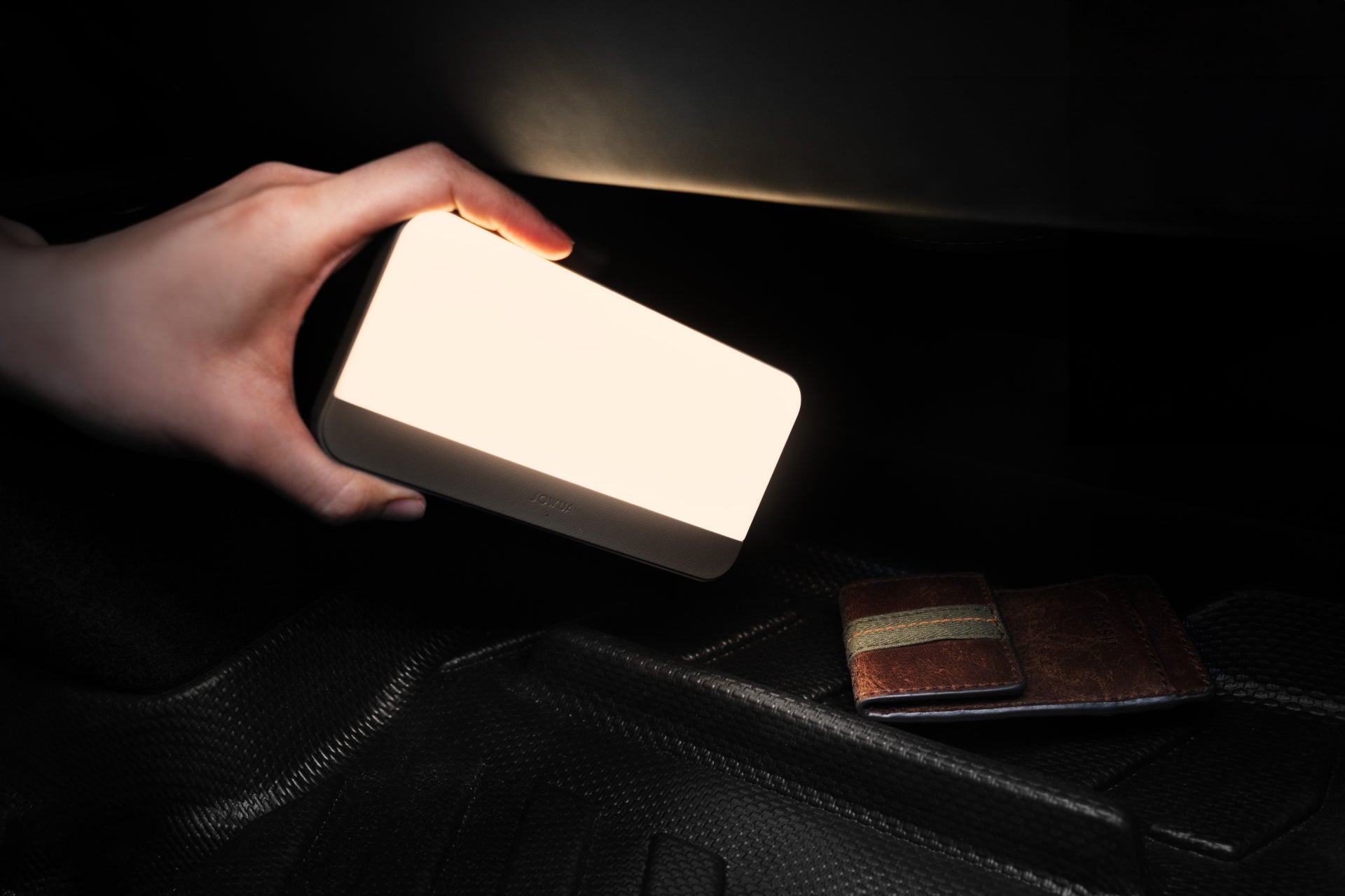 A hand holding a bright rectangular light source illuminates a dark space, revealing a brown leather wallet with a striped band resting beside the sleek JOWUA LED Center Console Combo - Tesla Reengineered Model 3 Highland (2024+) on the textured surface below.