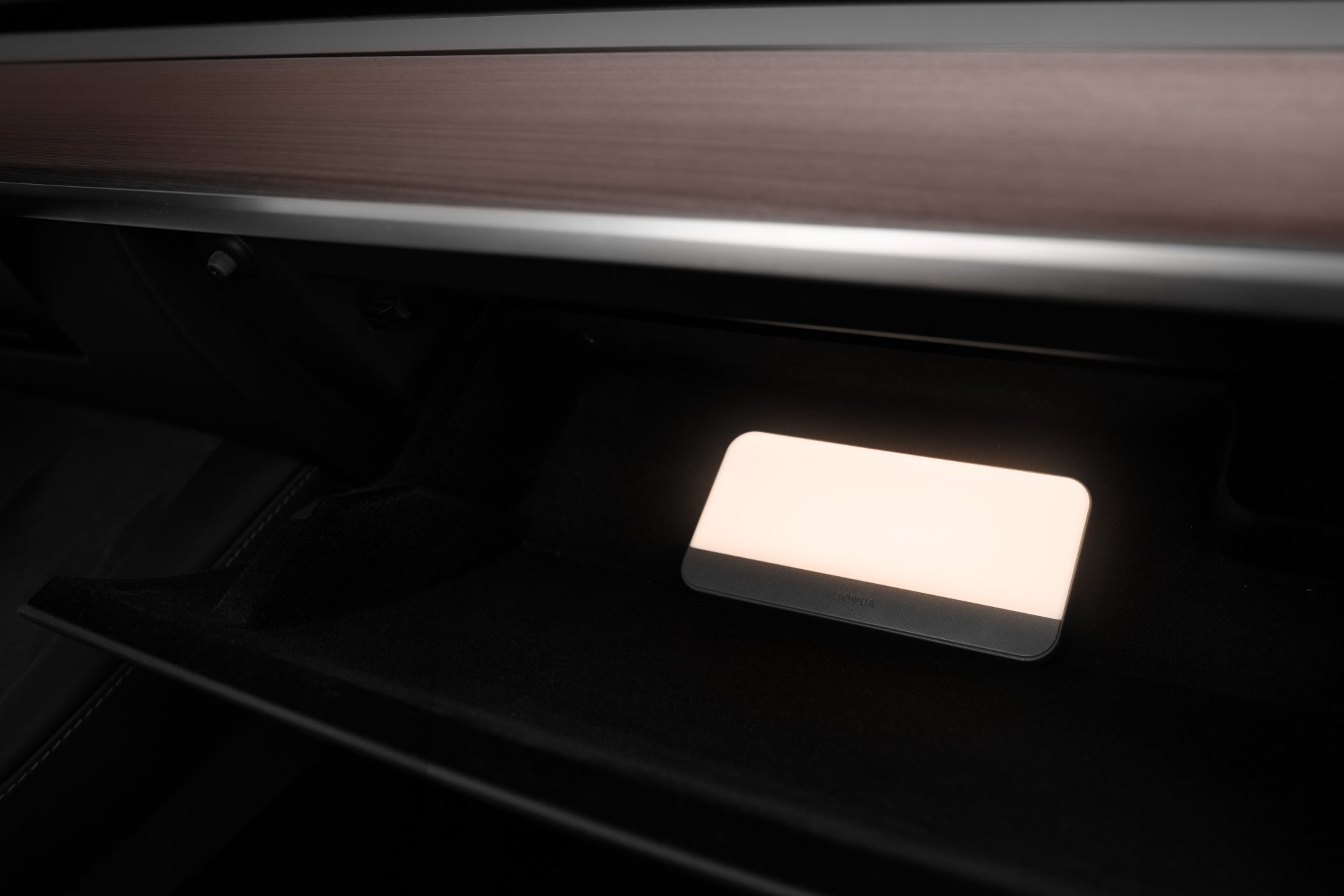 A dimly lit car interior reveals an open glove compartment illuminated by the soft glow of a rectangular light. The sleek wooden-finished dashboard is enhanced by the JOWUA LED Center Console Combo for Tesla Reengineered Model 3 Highland (2024+), adding both style and functionality with its premium silicone organizer.