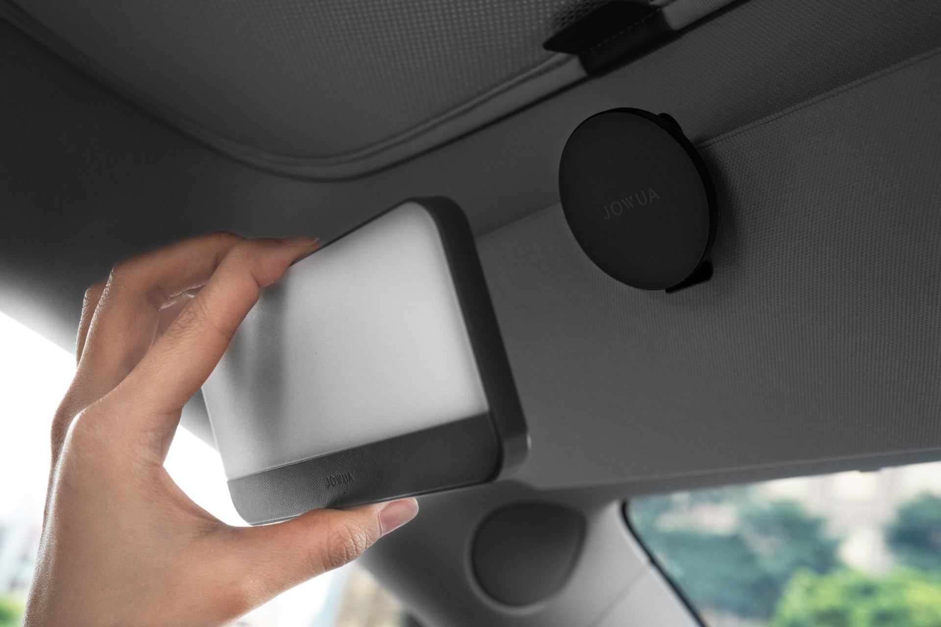 A person holds a rectangular device near a round magnetic mount on a car's interior roof. Both items, made from premium silicone, are branded with the name 