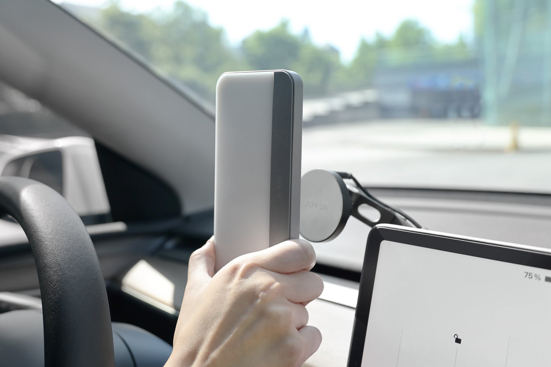 A person in a car is holding a smartphone in front of a mounted device on the dashboard. The steering wheel and part of a tablet screen, displaying a battery icon at 75%, are visible, while the JOWUA LED Center Console Combo for the Tesla Reengineered Model 3 Highland (2024+) is crafted from premium silicone and conveniently stores essentials nearby.