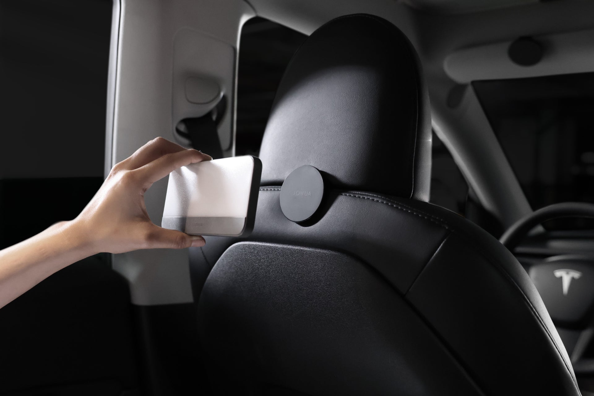 A person installs a wireless charging pad on the back of a car seat. The car interior, accented with a Tesla logo on the steering wheel, features the JOWUA LED Center Console Combo - Tesla Reengineered Model 3 Highland (2024+), which includes premium silicone organizers in the center console while securing the charging pad neatly on the headrest.