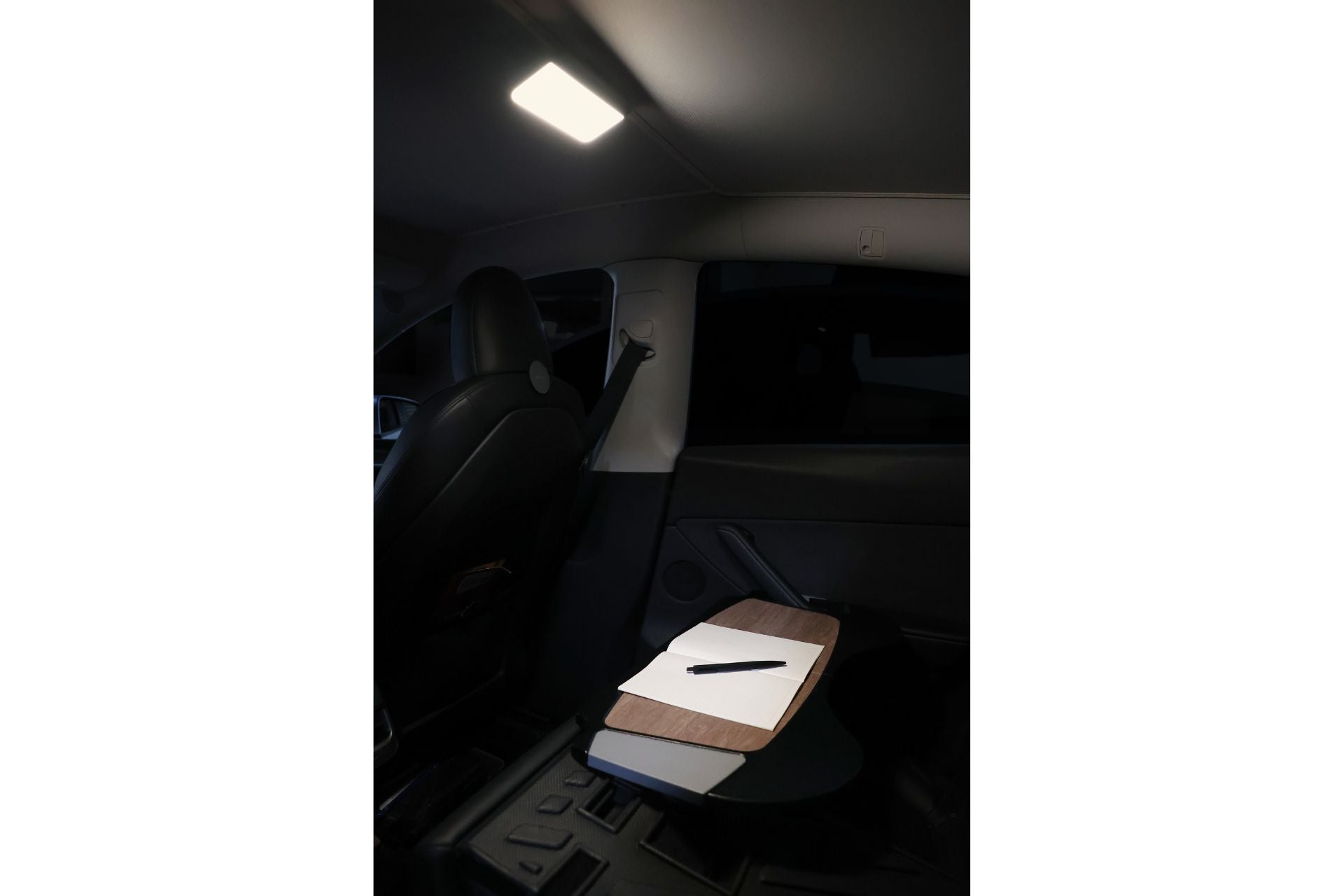 The image depicts the interior of a car with a light from the rear seat area illuminating the space. A folded table hosts a book, a closed notebook, and a pen. A JOWUA LED Center Console Combo for the Tesla Reengineered Model 3 Highland (2024+) keeps essentials organized in premium silicone, creating an efficient workspace within the vehicle.