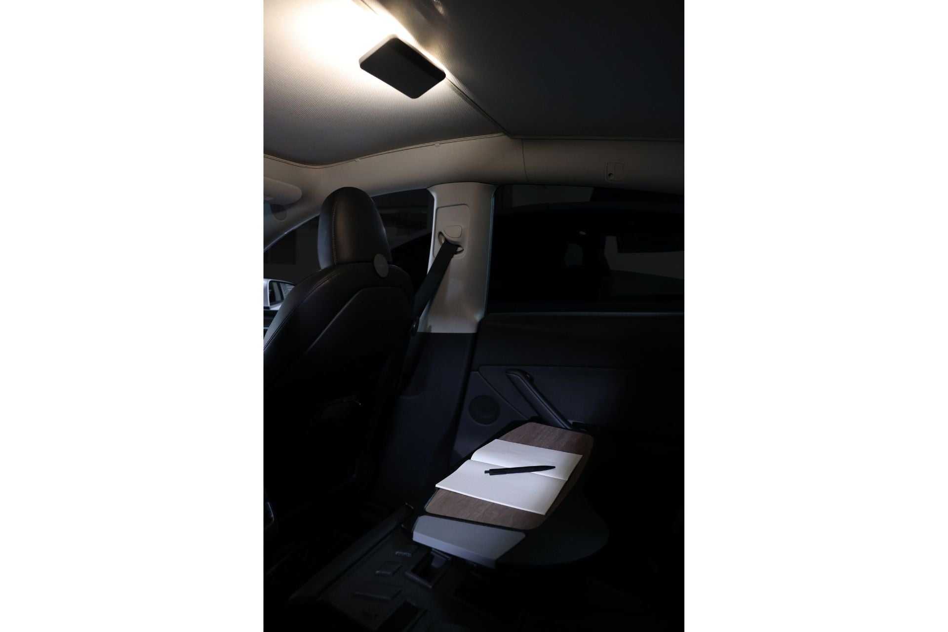 The interior of the car is softly illuminated by a ceiling light, highlighting a wooden folding table with a white surface and a pen on it in the backseat area. To the left, the driver's seat is visible. The setup is enhanced by JOWUA's LED Center Console Combo - Tesla Reengineered Model 3 Highland (2024+), crafted in premium silicone for added sophistication and functionality.