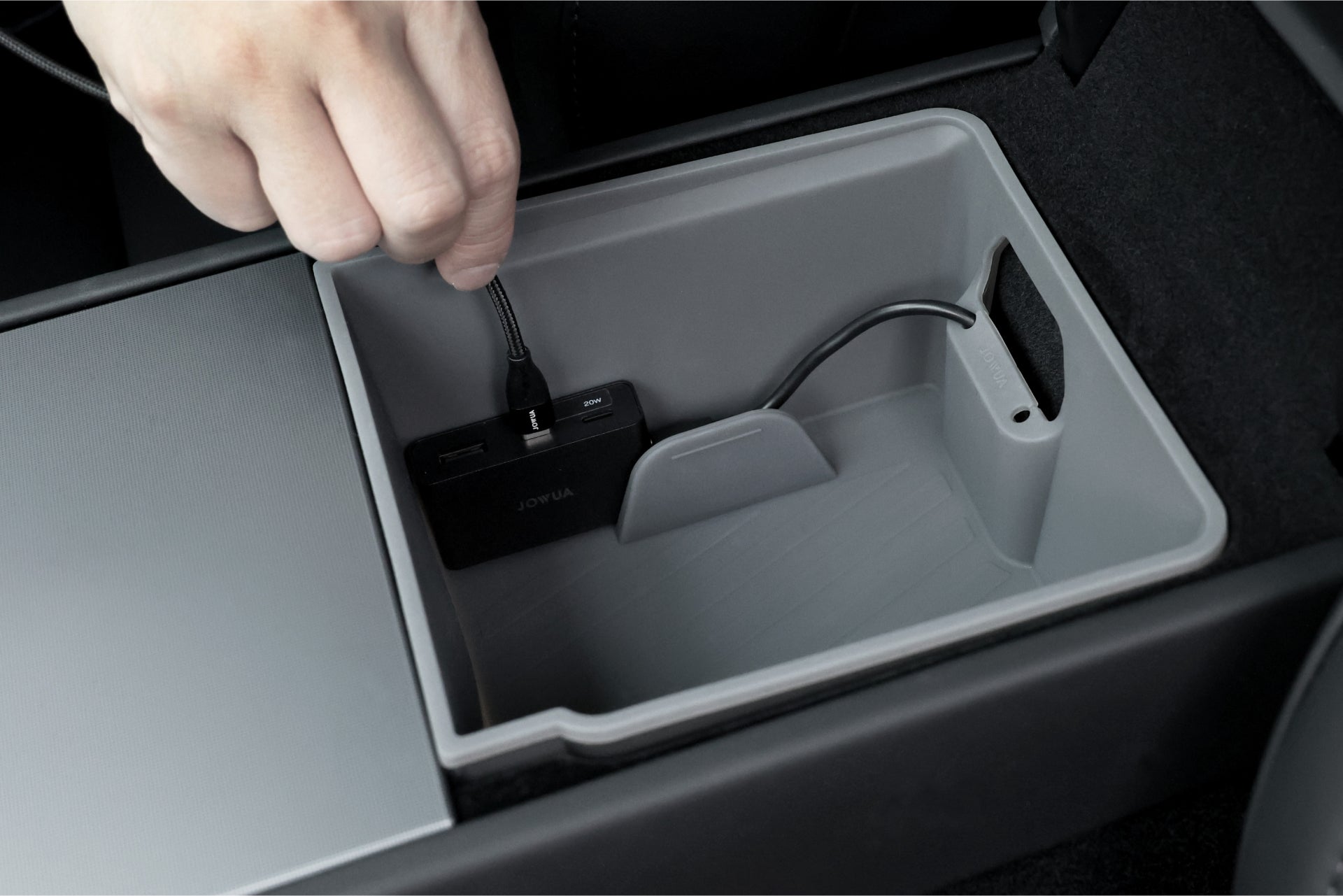 A hand connects a cable to the JOWUA Power HUB, highlighting its multi-device charging capability within the car's central console. The light gray compartment showcases a compact design, with USB-C power conversion and cables neatly organized.