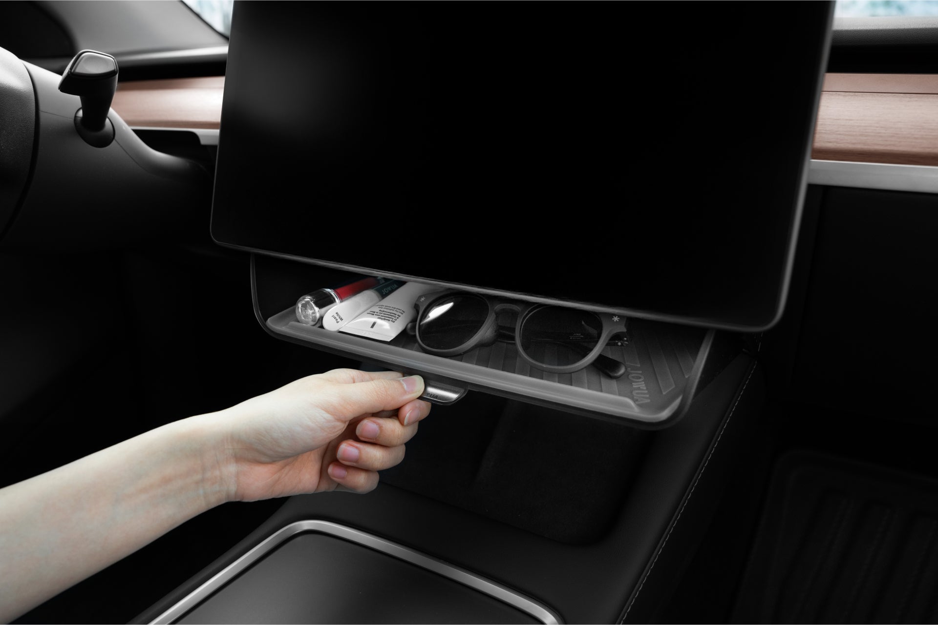 A person unveils the JOWUA Under Screen Invisible Station beneath the car's dashboard, revealing sunglasses, a pen, and keys secured by a magnetic snap. The sleek interior design with wood accents blends seamlessly with its sophisticated hidden integration.