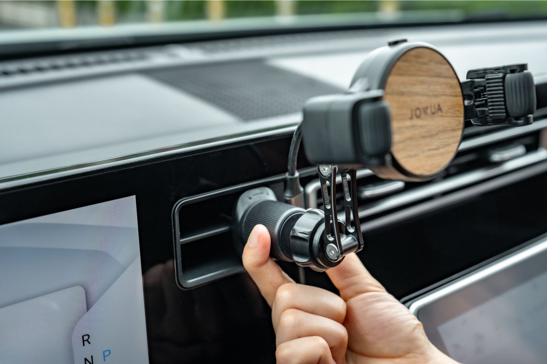 A hand adjusts a JOWUA Universal Air Vent Car Mount (Qi2 Universal Roller Mount), featuring a wooden back and Qi2 Wireless Charging, on a vehicle's air vent, with part of the digital display visible on the dashboard.