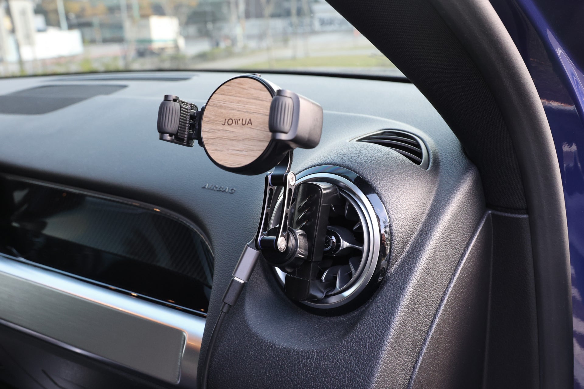 An elegant air vent car mount with adjustable clamp arms, featuring a wood-style design and the "JOWUA" brand. The dashboard and air vent controls are partially visible, showcasing the Universal Air Vent Car Mount (Qi2 Universal Roller Mount).