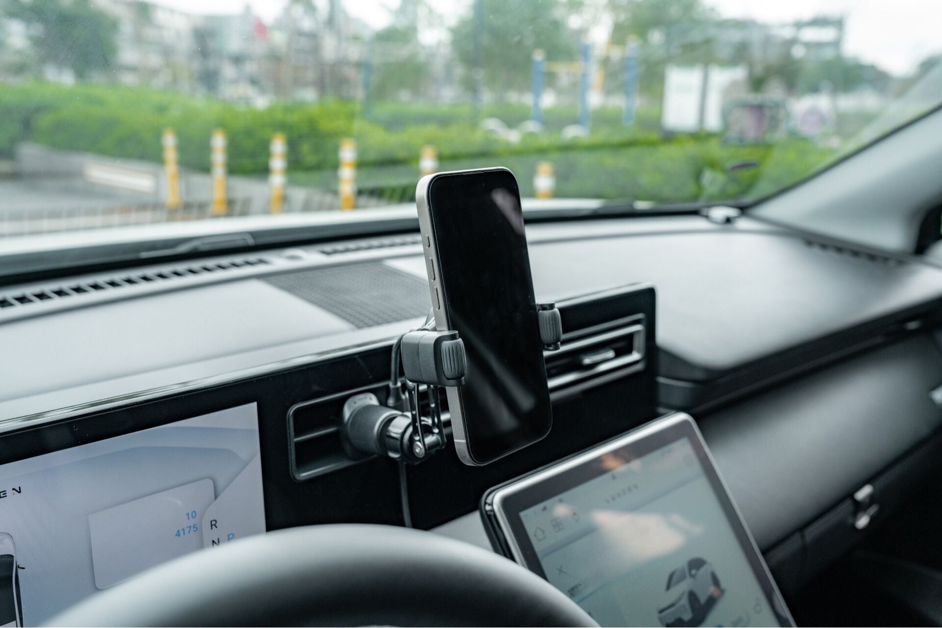 A JOWUA Universal Air Vent Car Mount (Qi2 Universal Roller Mount) is affixed to the car dashboard by the steering wheel. The securely mounted phone displays navigation and car settings, while green scenery through the windshield complements the tech with nature.