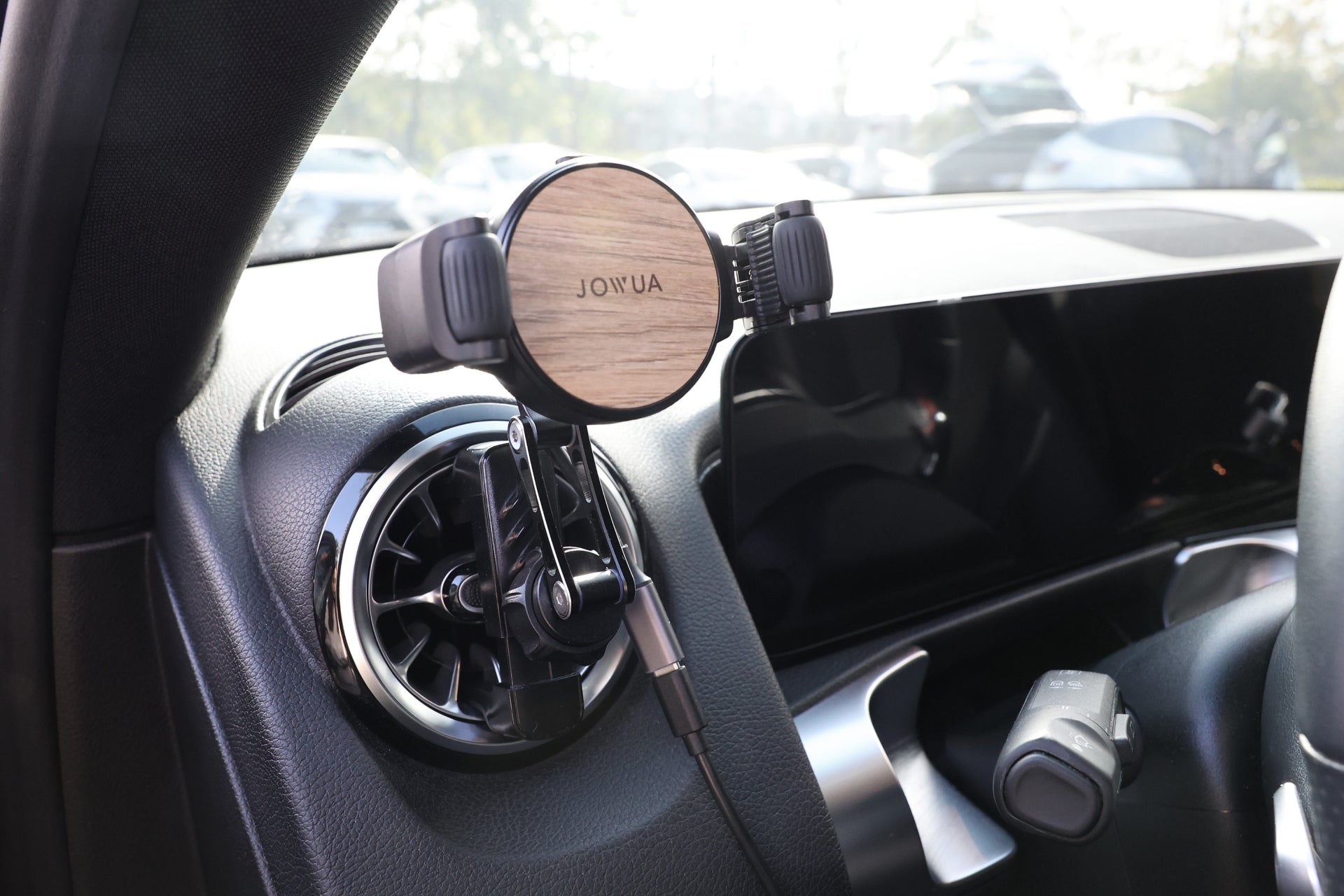 A sleek car interior with a modern dashboard showcases a JOWUA Universal Air Vent Car Mount. This elegant setup includes a touchscreen, gear selector, and the mount features Qi2 Vent Mount tech and Silicone Roller Clamps for secure placement, all bathed in sunlight.