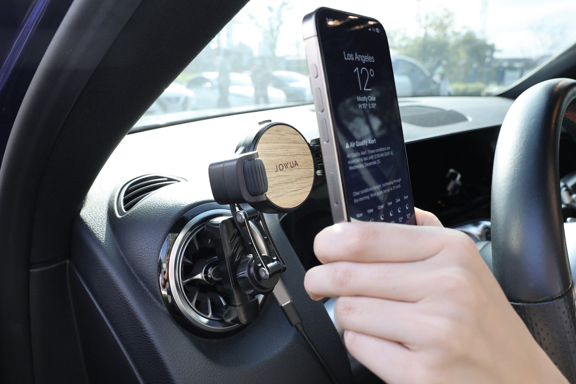 A person places a smartphone into a JOWUA Qi2 Universal Roller Mount with silicone clamps attached to an air vent. The phone screen shows a weather app with Los Angeles info while the steering wheel and car interior are partially visible.