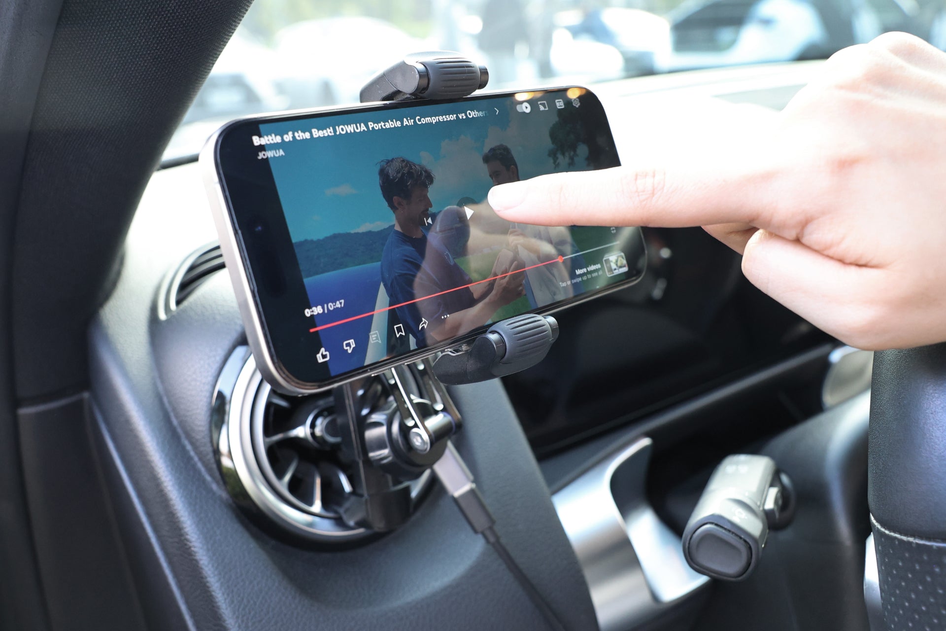 A person interacts with a smartphone, which is playing a video and held near the air vent by the JOWUA Universal Air Vent Car Mount (Qi2 Universal Roller Mount). Sunlight brightens the car interior, enhancing the sleek setup.