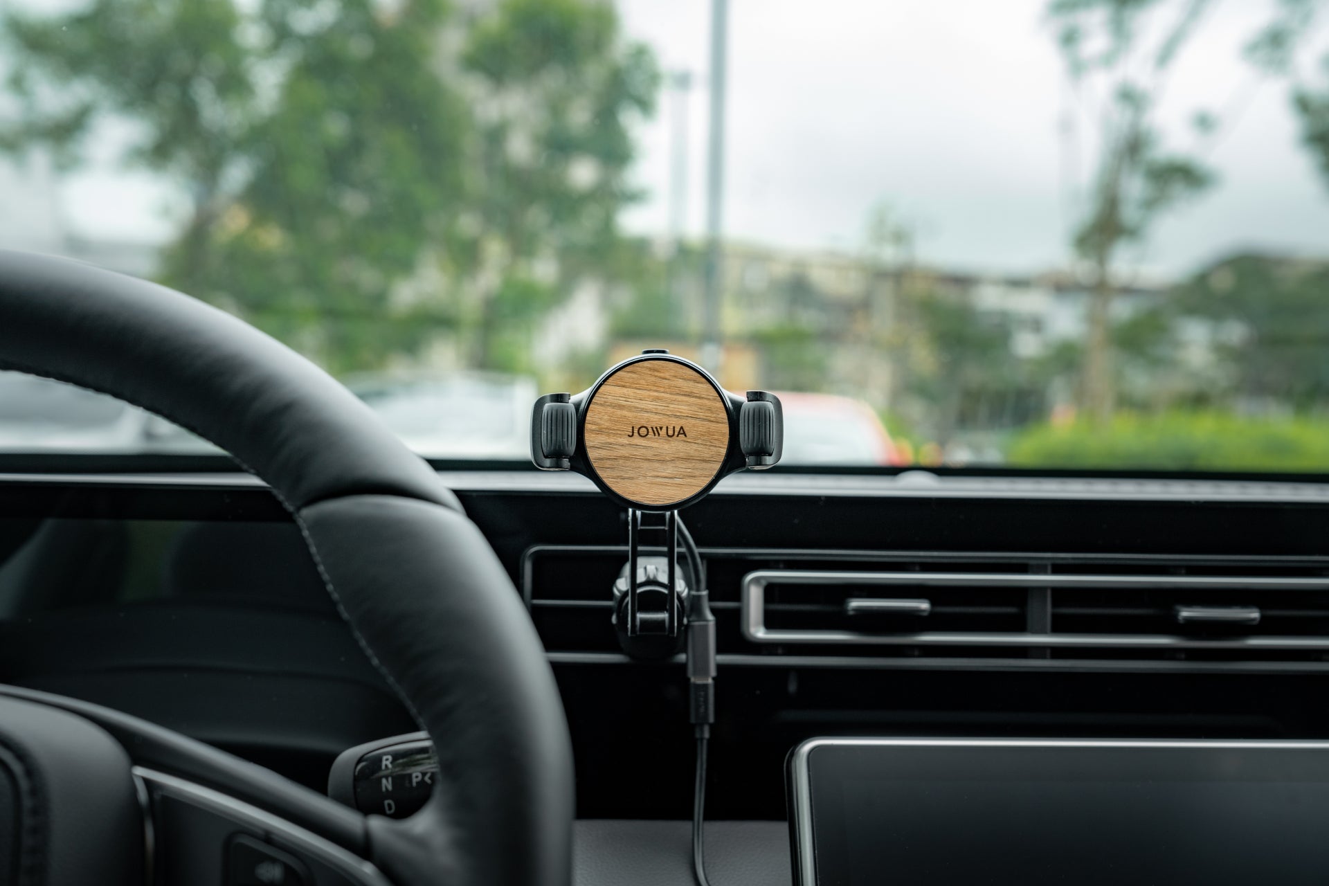 A stylish JOWUA wooden phone holder with adjustable clamp arms, the Universal Air Vent Car Mount, is mounted on the air vent. It complements the steering wheel and gear shift as trees and buildings blur softly through the windshield.