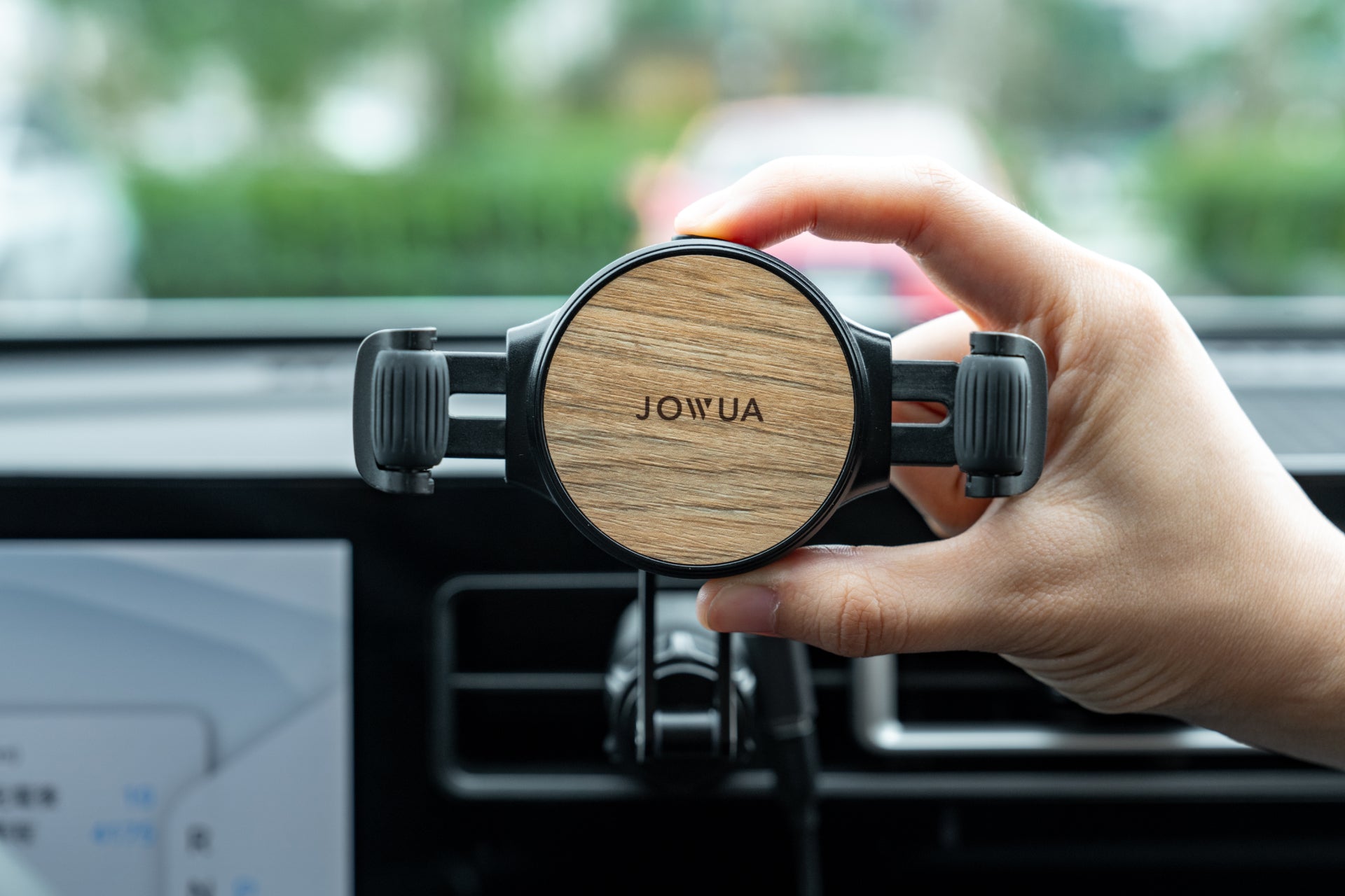 A hand adjusts a wooden Universal Air Vent Car Mount (Qi2 Universal Roller Mount) by JOWUA, featuring silicone roller clamps on the dashboard vent. The car's interior and touchscreen are visible with blurred greenery and a parked car in the background.