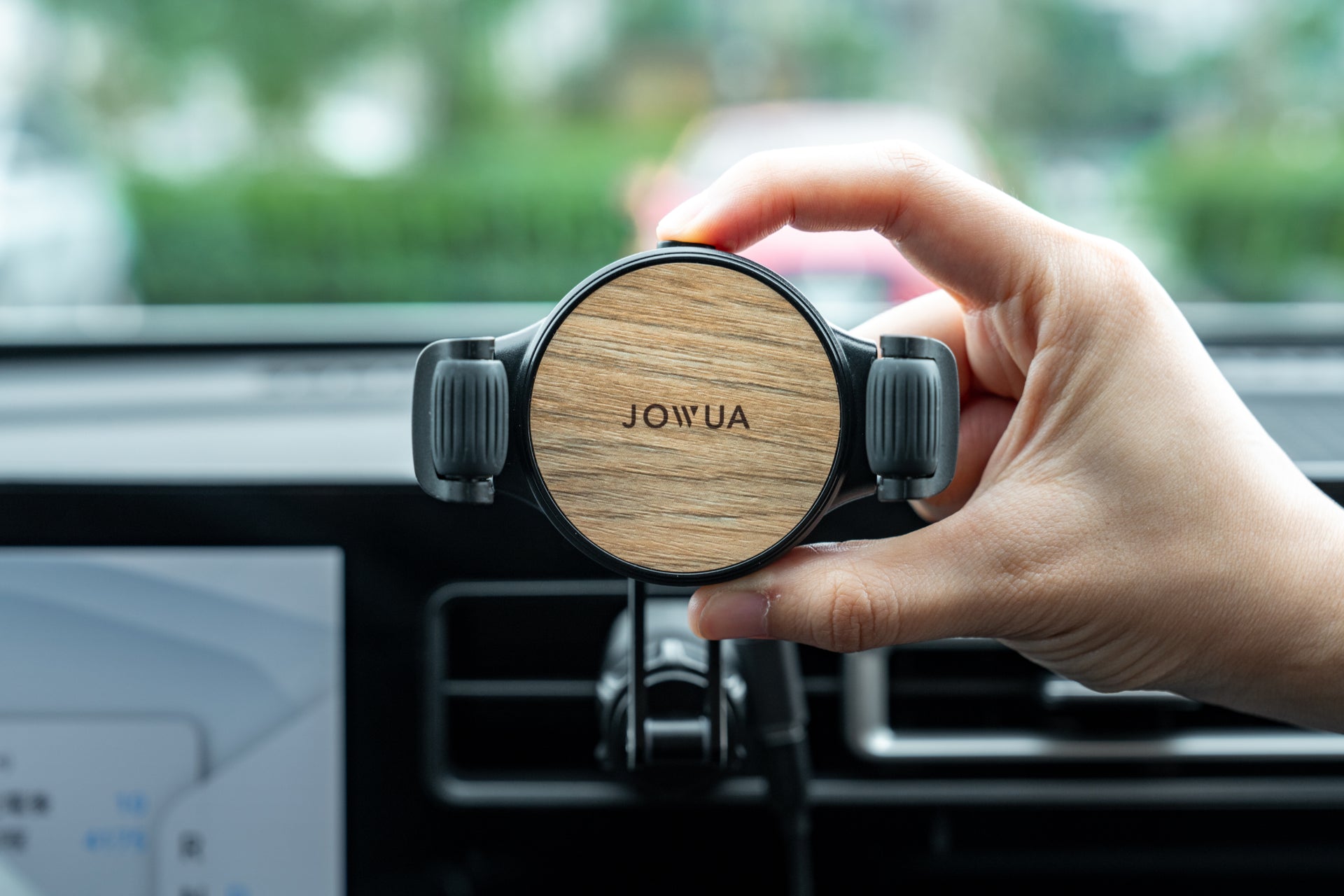 A hand holding JOWUA's Universal Air Vent Car Mount (Qi2 Universal Roller Mount), featuring adjustable clamp arms and Qi2 wireless charging, inside a car with dashboard details and a blurry view of greenery and another vehicle visible outside the window.