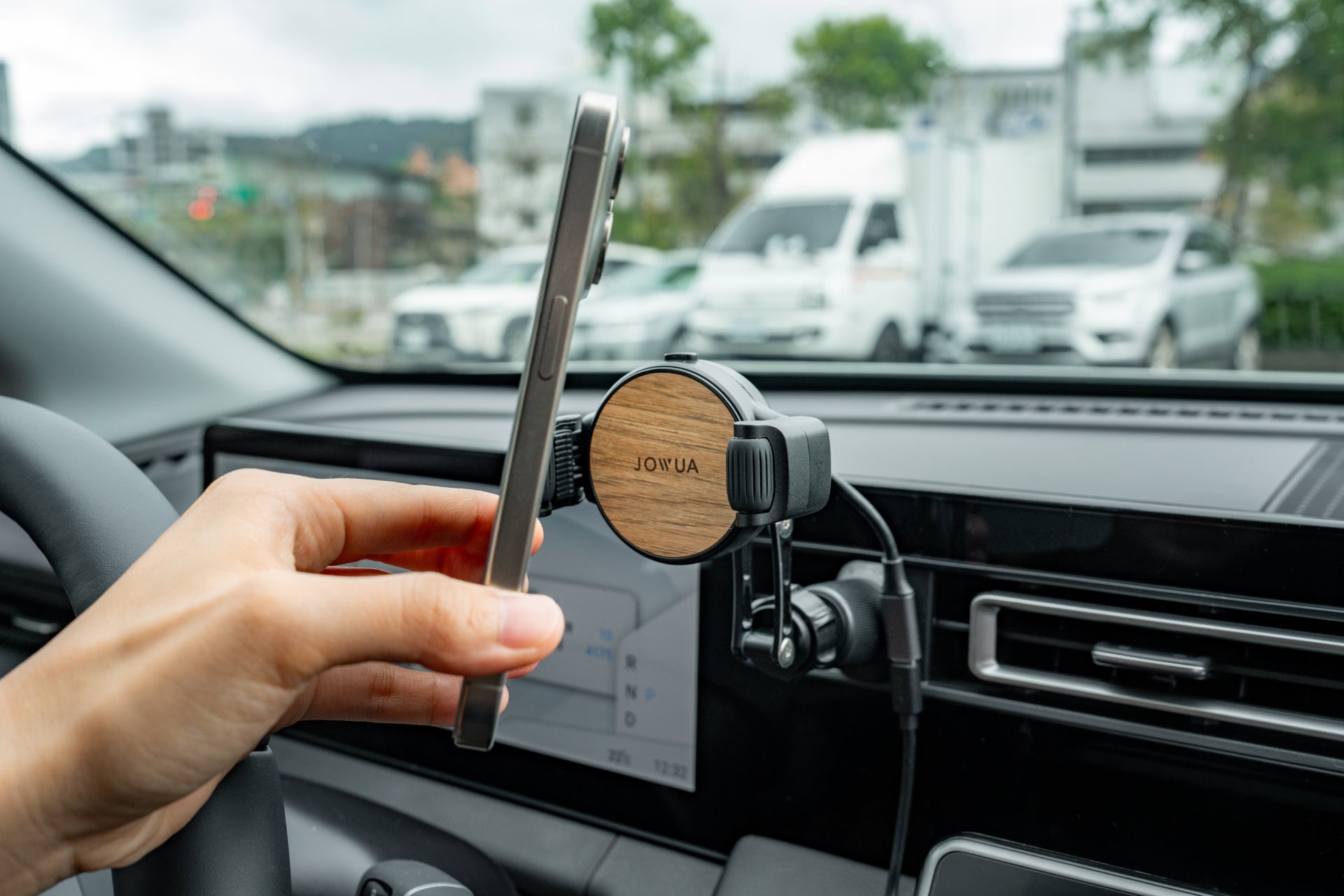 An individual secures their smartphone to the JOWUA Universal Air Vent Car Mount (Qi2 Universal Roller Mount) using its silicone roller clamps inside a vehicle, with a blurred backdrop of parked cars and lush greenery visible outside.