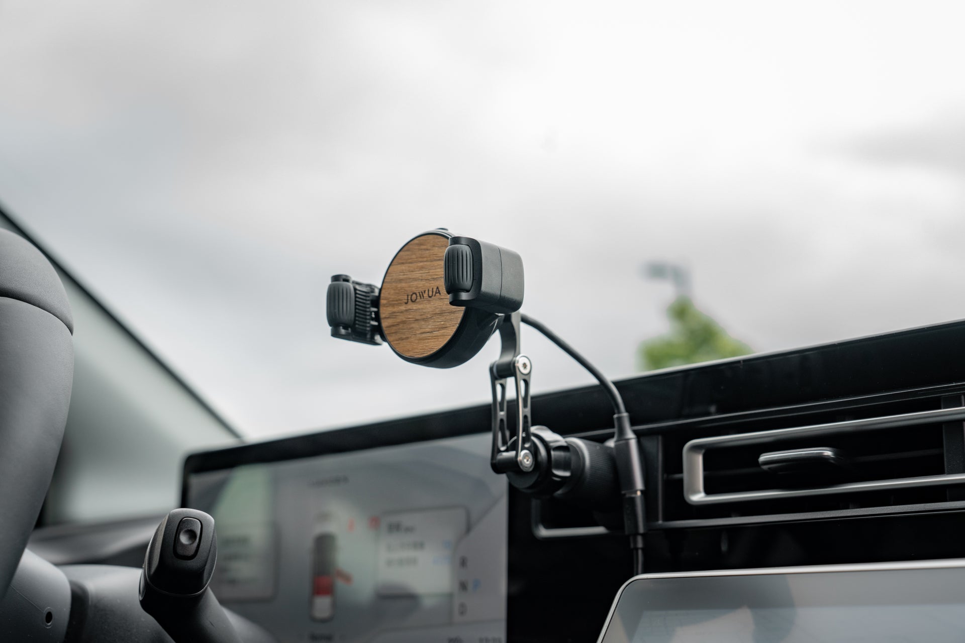 A JOWUA Universal Air Vent Car Mount with a wooden backplate and adjustable clamp arms is attached near the steering wheel. It features Qi2 wireless charging and an adjustable arm, visible alongside parts of the steering wheel and infotainment system.