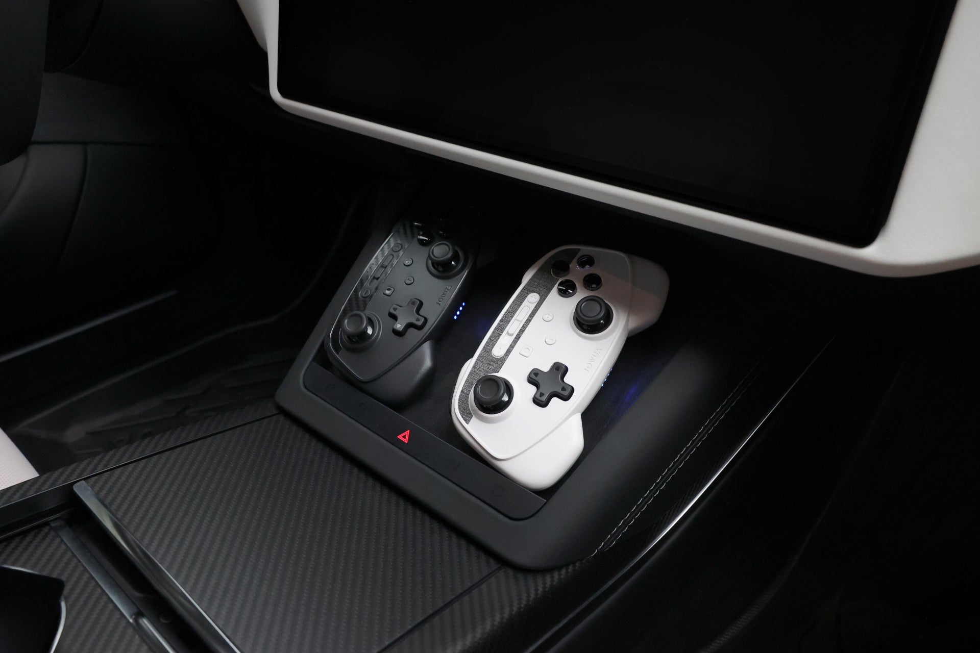 Inside a car interior, two JOWUA Wireless Tesla Game Controllers from the Model S/3/3HL/X/Y/Cyber series rest in the central console's charging dock. These black and white controllers, crafted for Bluetooth Connect, are seamlessly integrated into a sleek and modern design featuring carbon fiber accents reminiscent of a Tesla Steam interface.