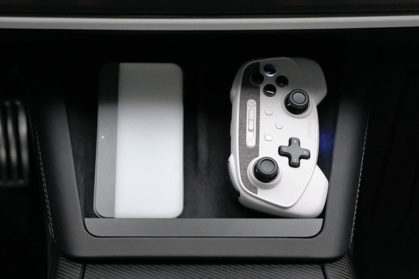 A pair of elegant Wireless Tesla Game Controllers from JOWUA, featuring a mix of white and black buttons, rests on a tray adjacent to a slim rectangular device within the vehicle's interior. These controllers offer seamless Bluetooth Connect. The backdrop showcases a textured surface and vehicle pedals, indicating Tesla Steam integration for an enhanced driving experience.