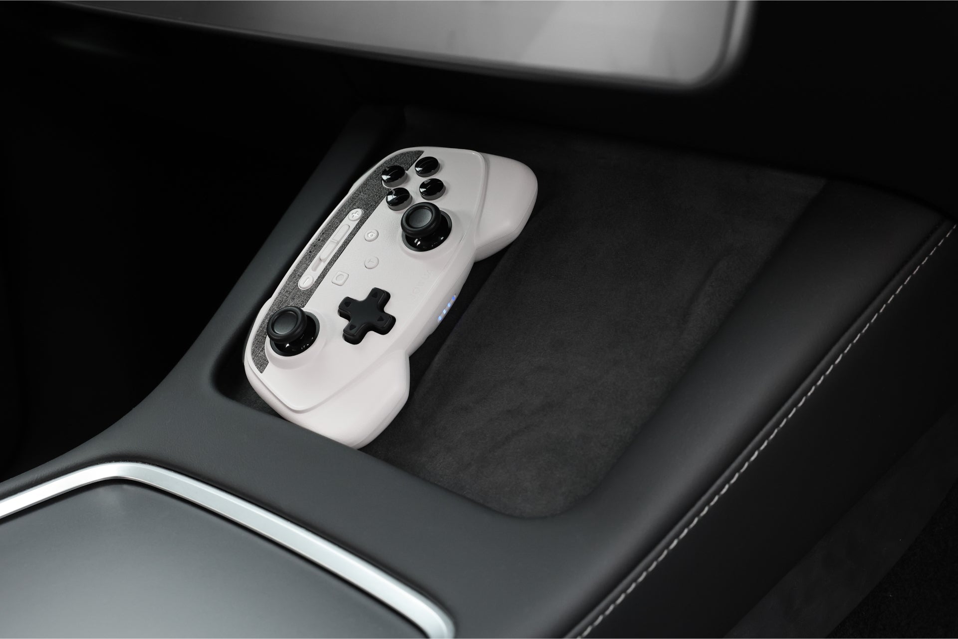 A JOWUA Wireless Tesla Game Controller from the Model S/3/3HL/X/Y/Cyber Combo, featuring a white design with black buttons, rests in a car's center console compartment. The controller connects effortlessly via Bluetooth Connect. The car's sleek interior, highlighted by dark upholstery and stitching detail, perfectly complements this modern tech setup.