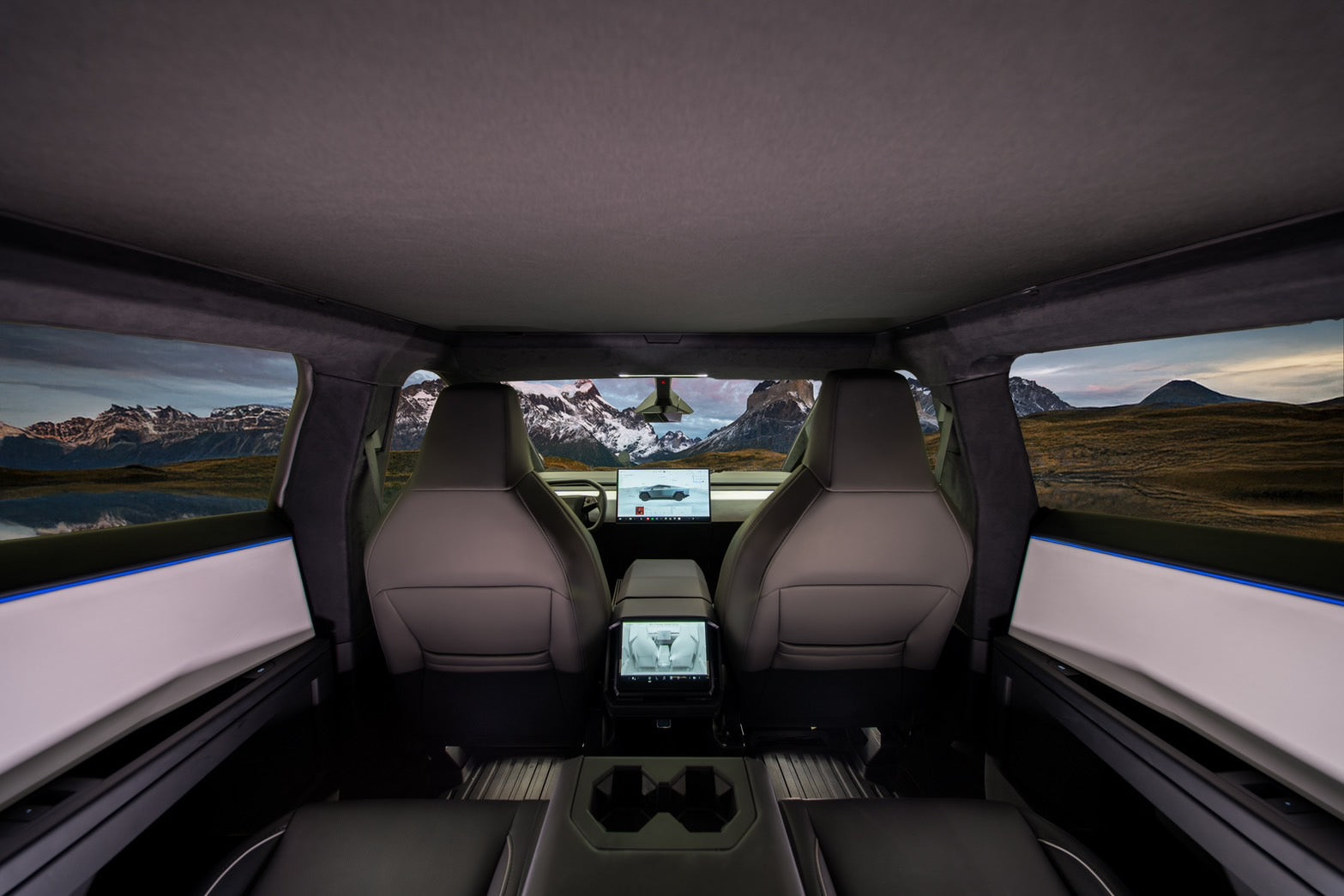 A view from inside a Cybertruck showcases the lightweight mesh fabric sunshade on the two front seats and a dashboard adorned with digital displays. The expansive windows, featuring JOWUA's Cybertruck Glass Roof Sunshade, offer UV protection while revealing a picturesque landscape of mountains and cloudy skies in the backdrop.