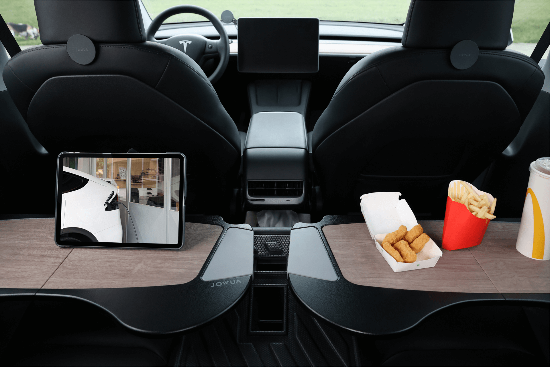 The interior of a Tesla Model S is adorned with premium materials, including a sleek wooden dashboard and two black front seats. A tablet on the left showcases a vehicle image, while in the center, there's a JOWUA Foldable Car Tray for Tesla holding a small carton of fries, sauce packet, and drink.