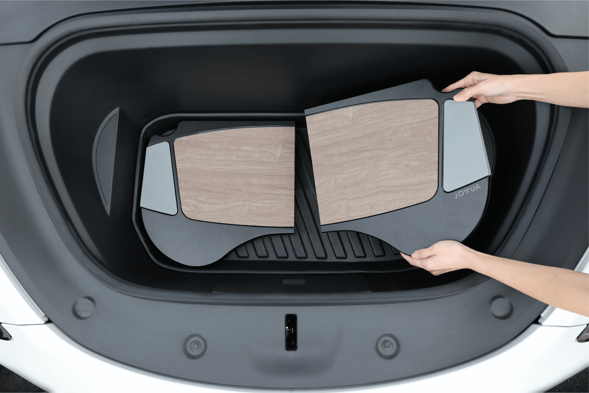 A person installs two JOWUA Foldable Car Trays featuring wood-like panels into the front trunk of their Tesla Model S/3/X/Y, highlighting these premium accessories. The white car body around the trunk accentuates the black lining inside, underscoring its use of top-quality materials.