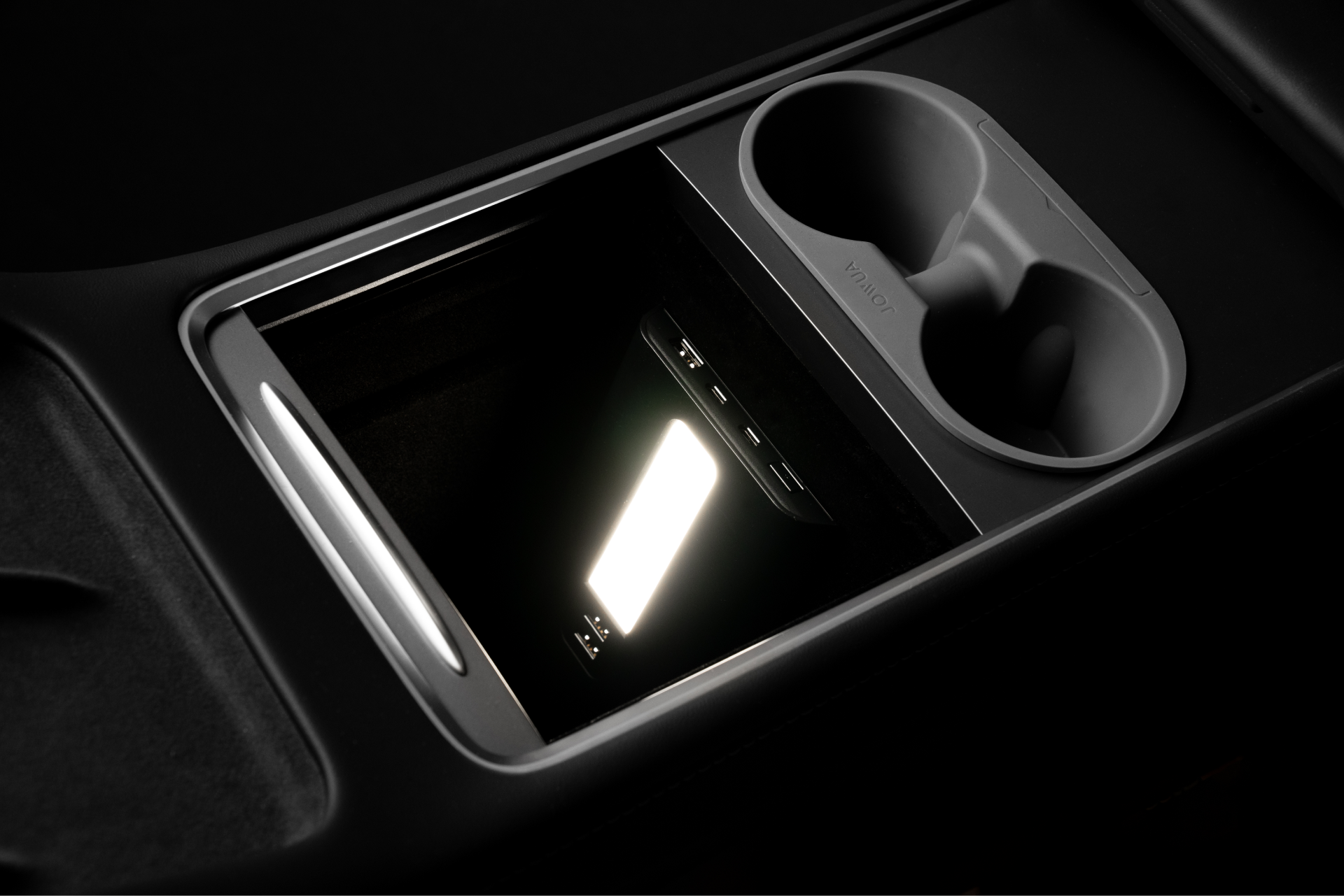 In the dimly lit interior of a Tesla Model 3, a smartphone lies on the center console next to two cup holders. The sleek design is emphasized by the soft glow from JOWUA's USB Hub for Tesla Model 3/Y, equipped with dual USB-C ports providing 45W power and an LED warm light to keep all your devices charged throughout your journey.