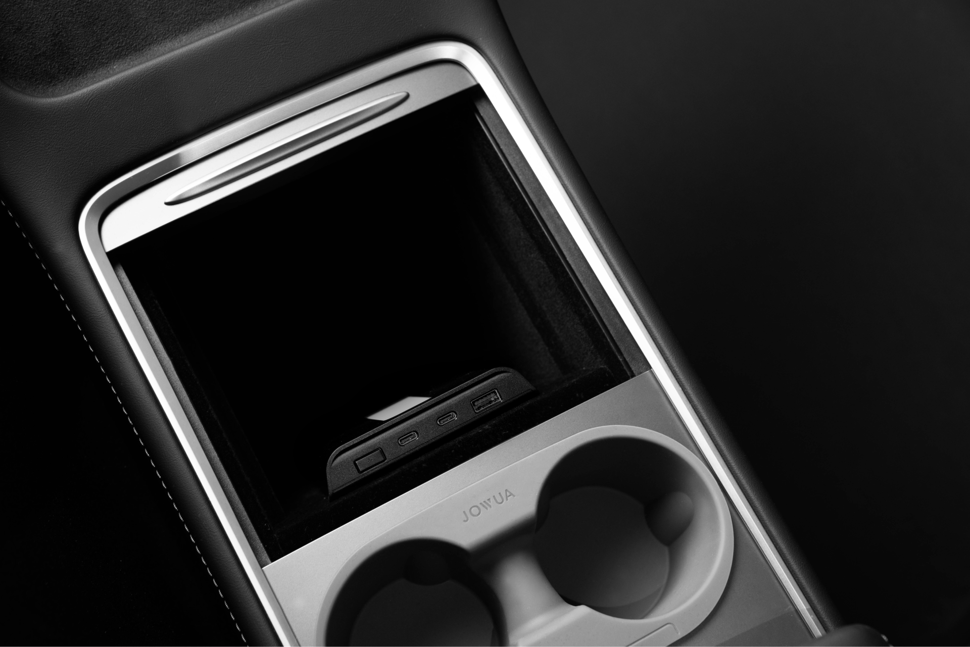 Black and white photo of a Tesla Model 3's center console showcasing a storage compartment with a remote control inside, two cup holders, and the JOWUA USB Hub for Tesla Model 3/Y featuring Dual USB-C 45W ports with LED Warm Light. The sleek, modern design is accentuated by the contrast of light and dark tones.