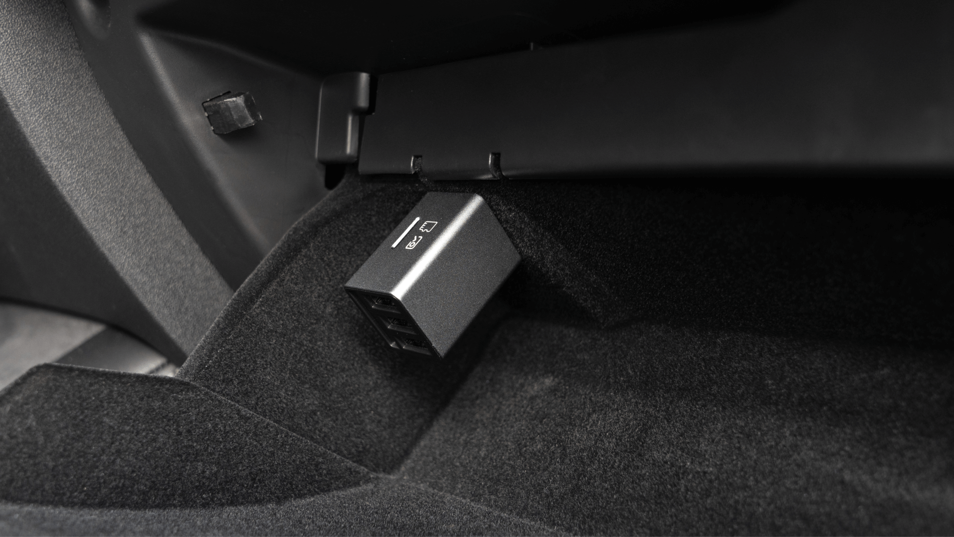 A black 3 Port USB HUB with DashCam Reader by JOWUA is plugged into a vehicle's port beneath the dashboard, seamlessly integrating with the HiSpeed USB connection. The interior features dark carpeting and a glimpse of the glove compartment.